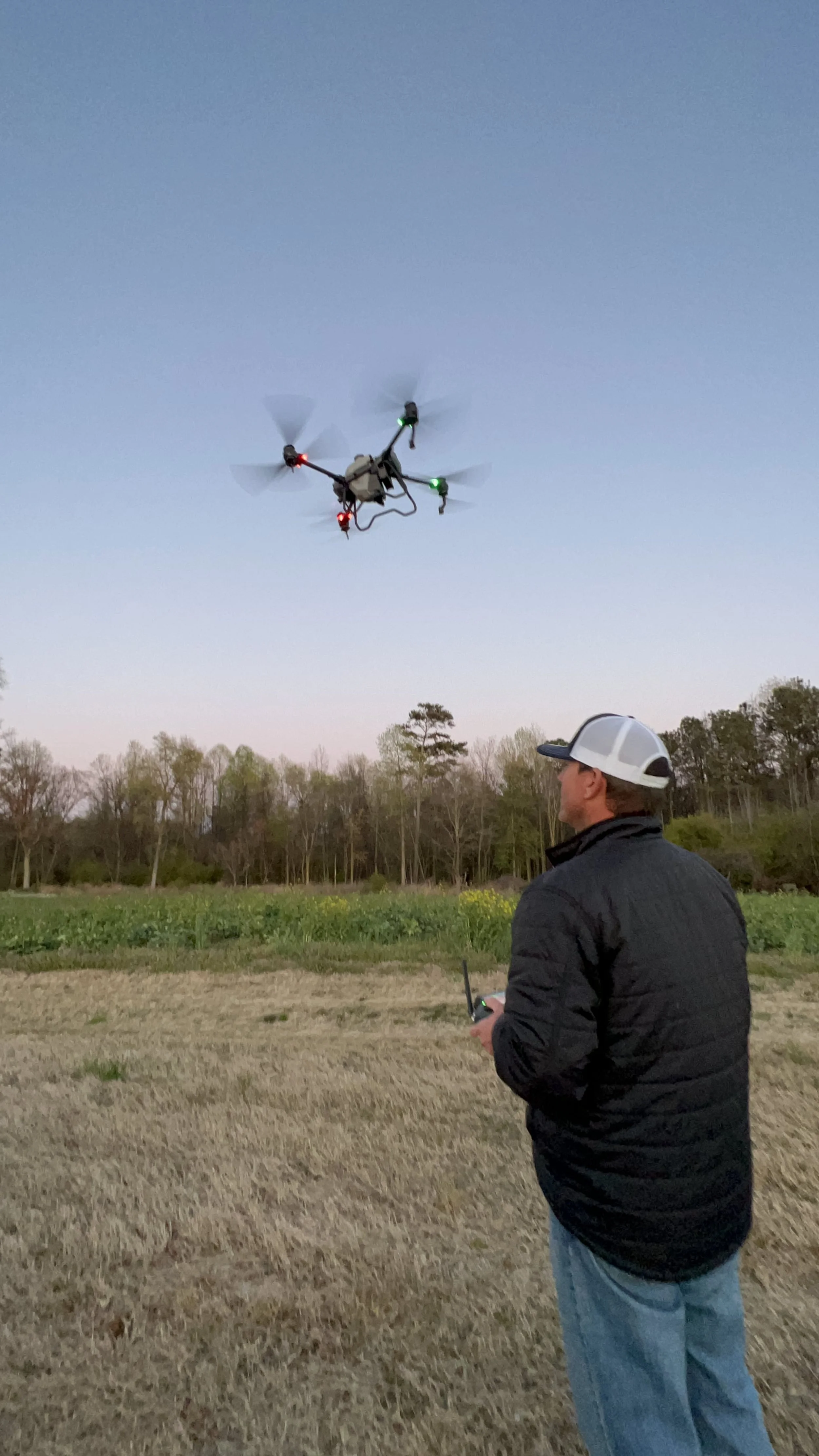 A man wearing a black jacket and a backward baseball cap controlling a drone with a remote in a field at sunset, with trees in the background.