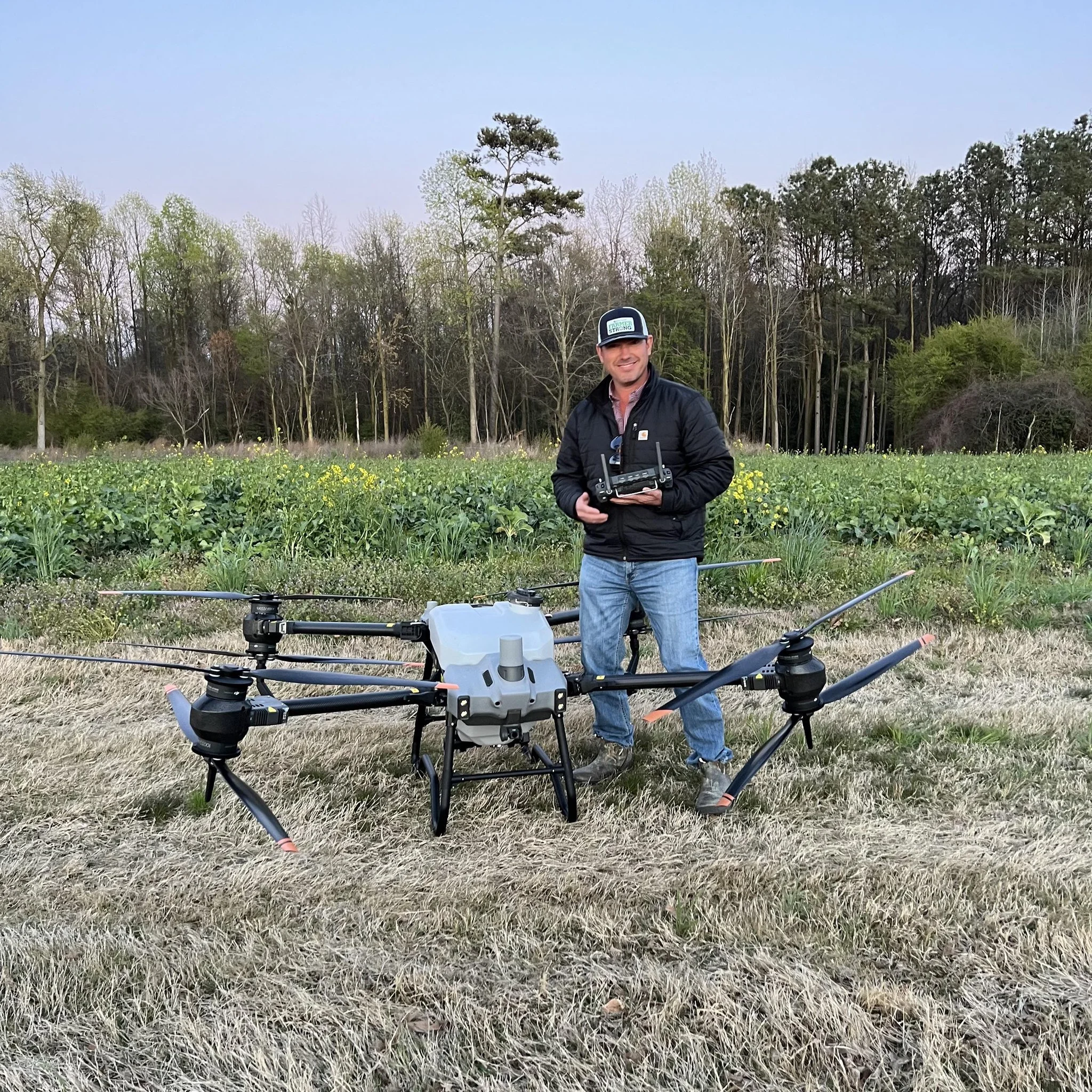 A man standing in a field holding a drone remote controller, with a large quadcopter drone on the ground in front of him, outdoors with trees and greenery in the background.