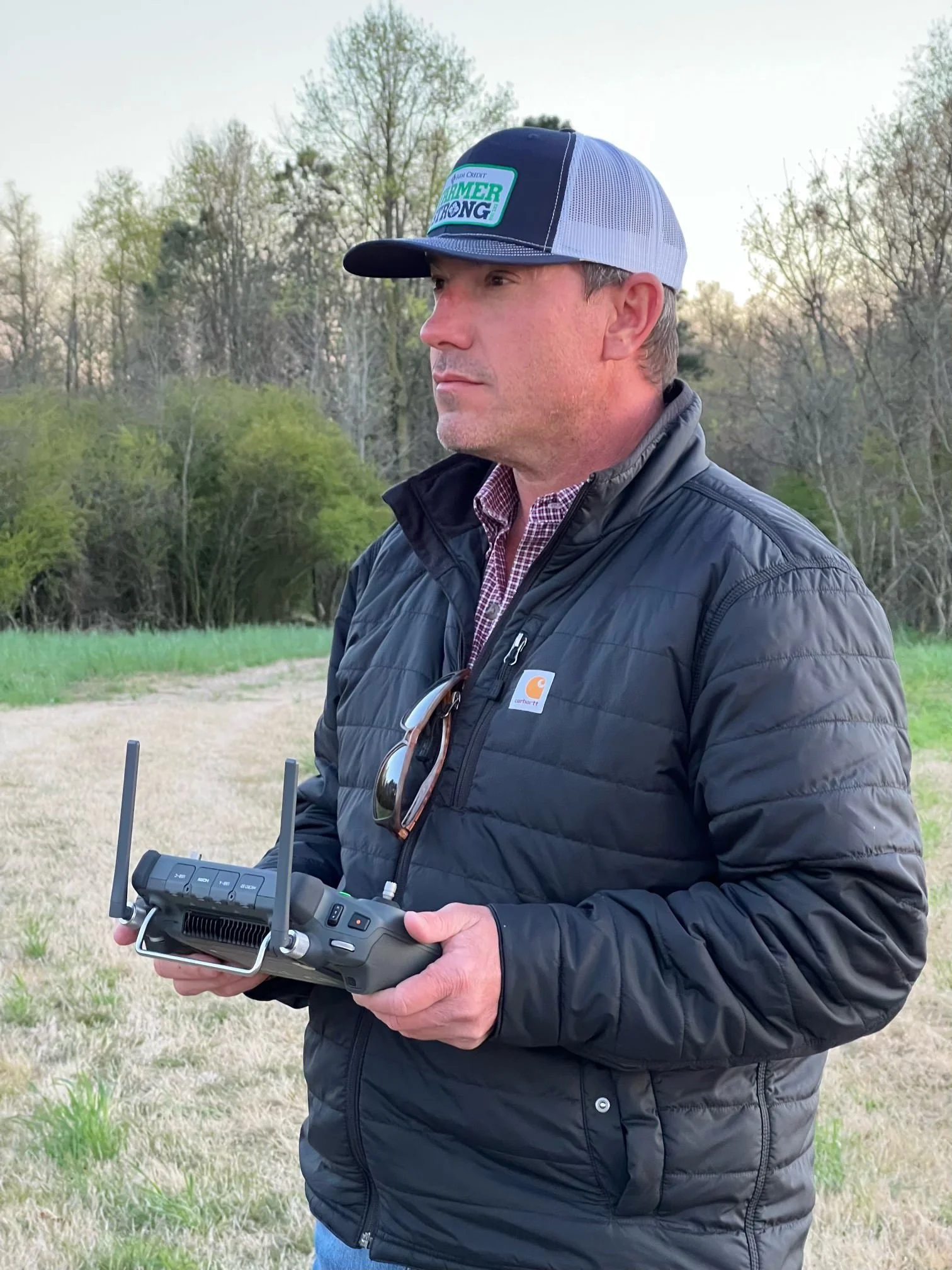 A man in a black puffer jacket holding a drone controller with antennas outdoors in a grassy area with trees in the background, wearing a baseball cap.