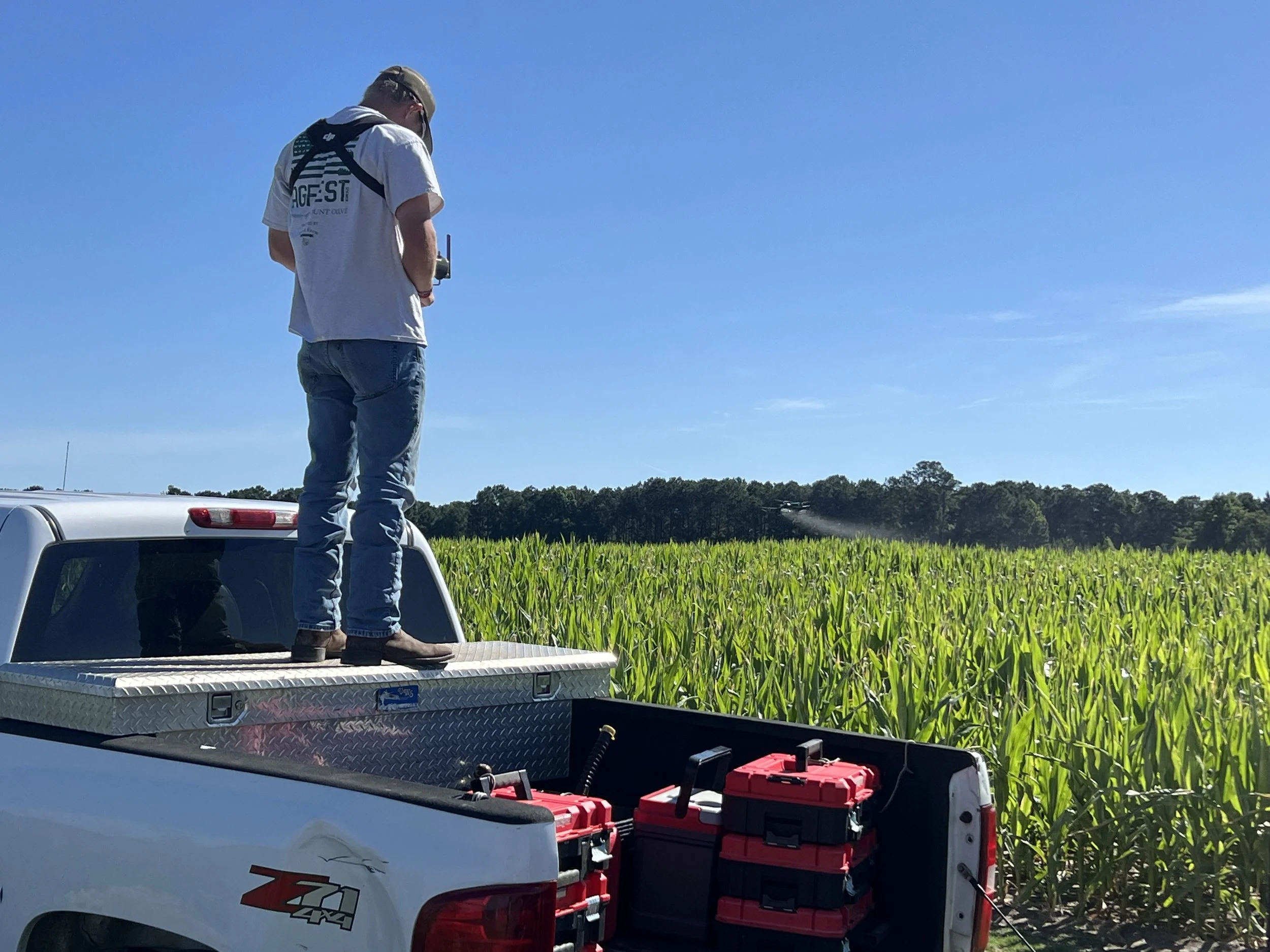A man standing on the back of a white pickup truck in a field of tall green crops, monitoring a device he is holding, with a clear blue sky overhead.