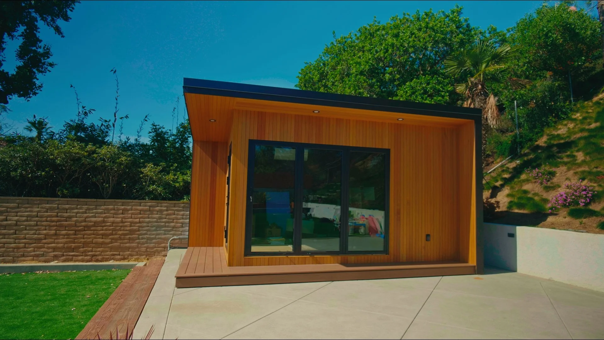 Small modern house with wooden exterior, black sliding glass doors, and a flat roof, set against a hillside with green trees and flowering plants.