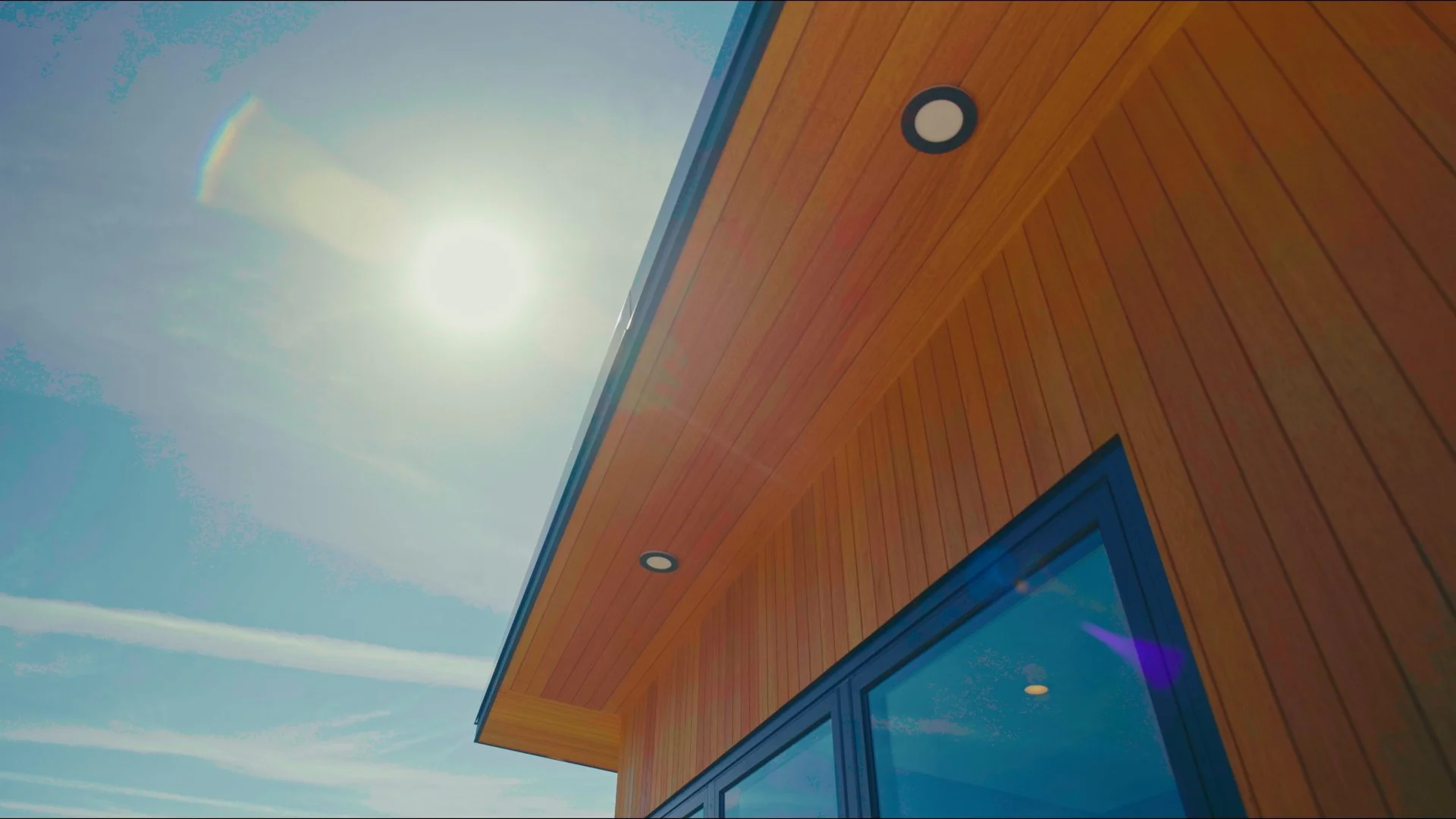 The image shows a bright sun shining in a clear blue sky, with a wooden building in the foreground. The building has a modern design with large glass windows and a wooden exterior.