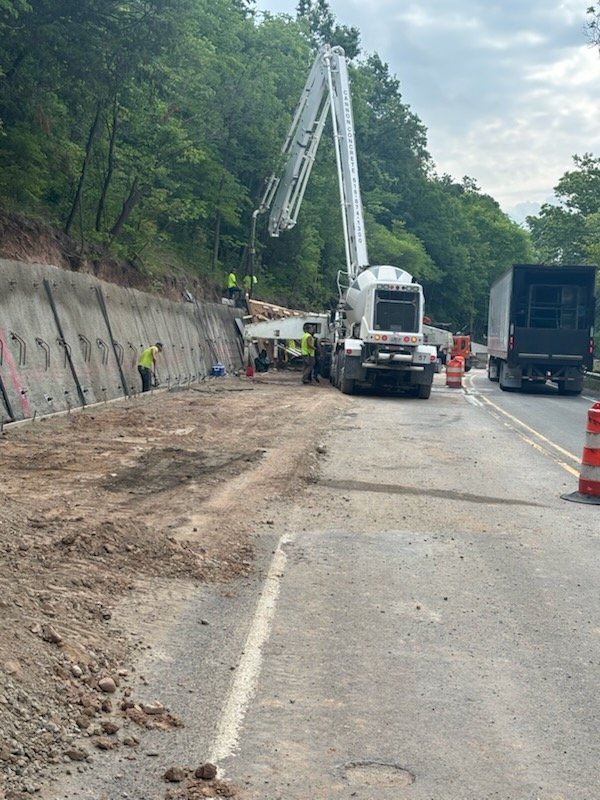 Road construction with workers and a crane on the side of a highway surrounded by trees.