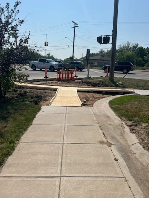 Sidewalk with a ramp, construction cones, and barriers, leading to a crosswalk at an intersection with traffic and utility poles.