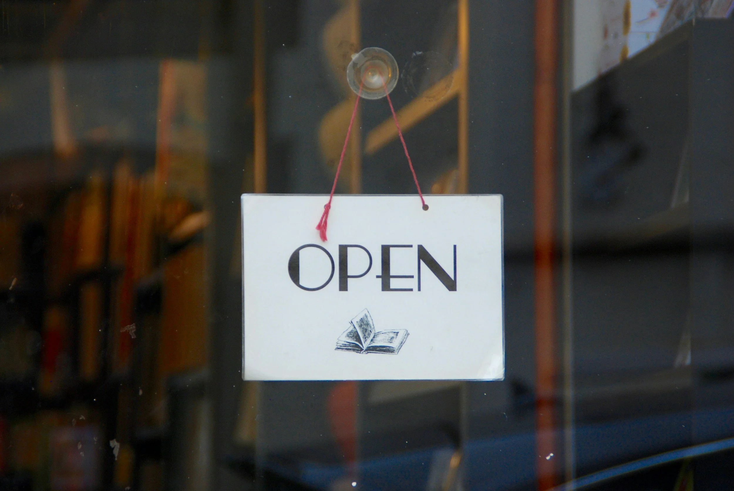 A white sign with the word "OPEN" and an illustration of an open book, hanging on a glass door.