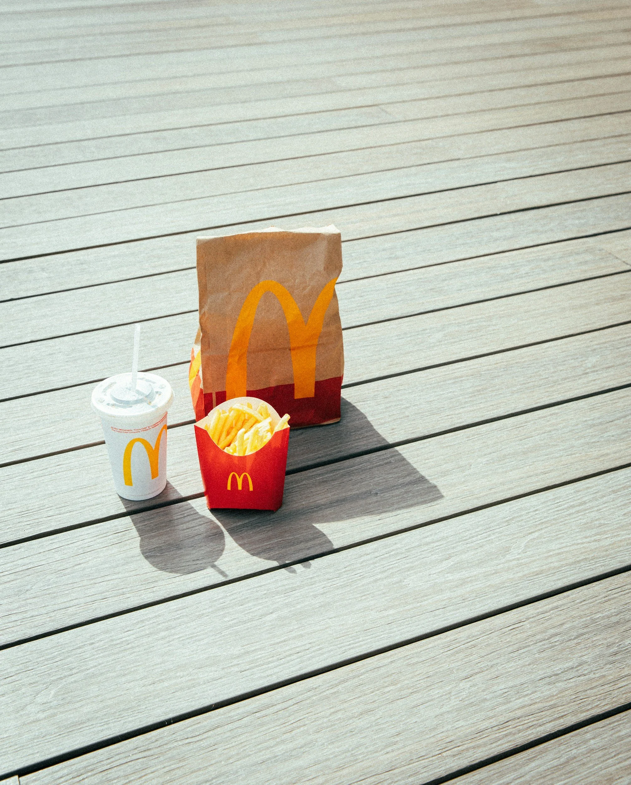 McDonald's french fry container, soft drink cup, and paper bag on a wooden deck.