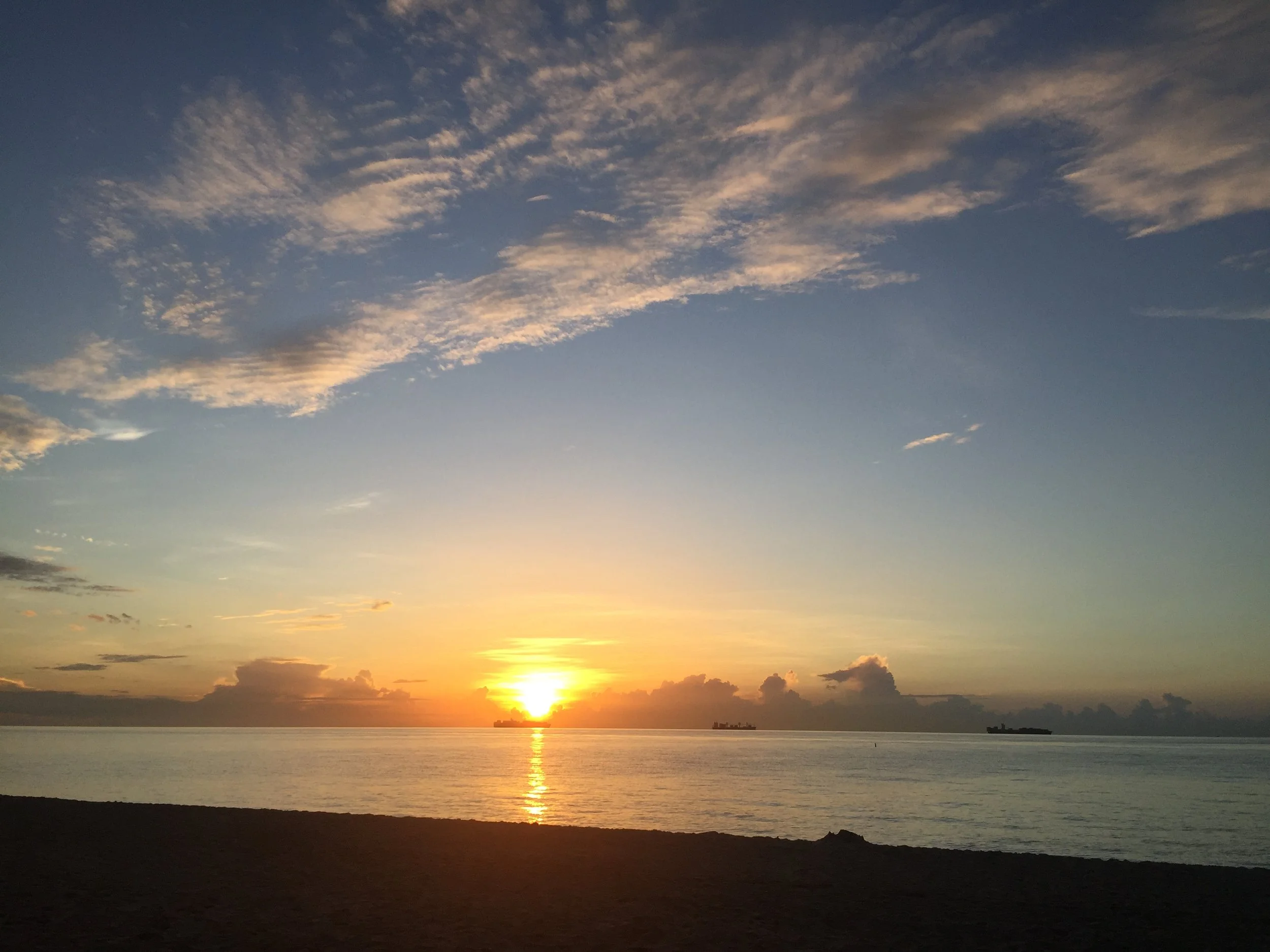 Sunset over the ocean with a few ships on the horizon and scattered clouds in the sky.