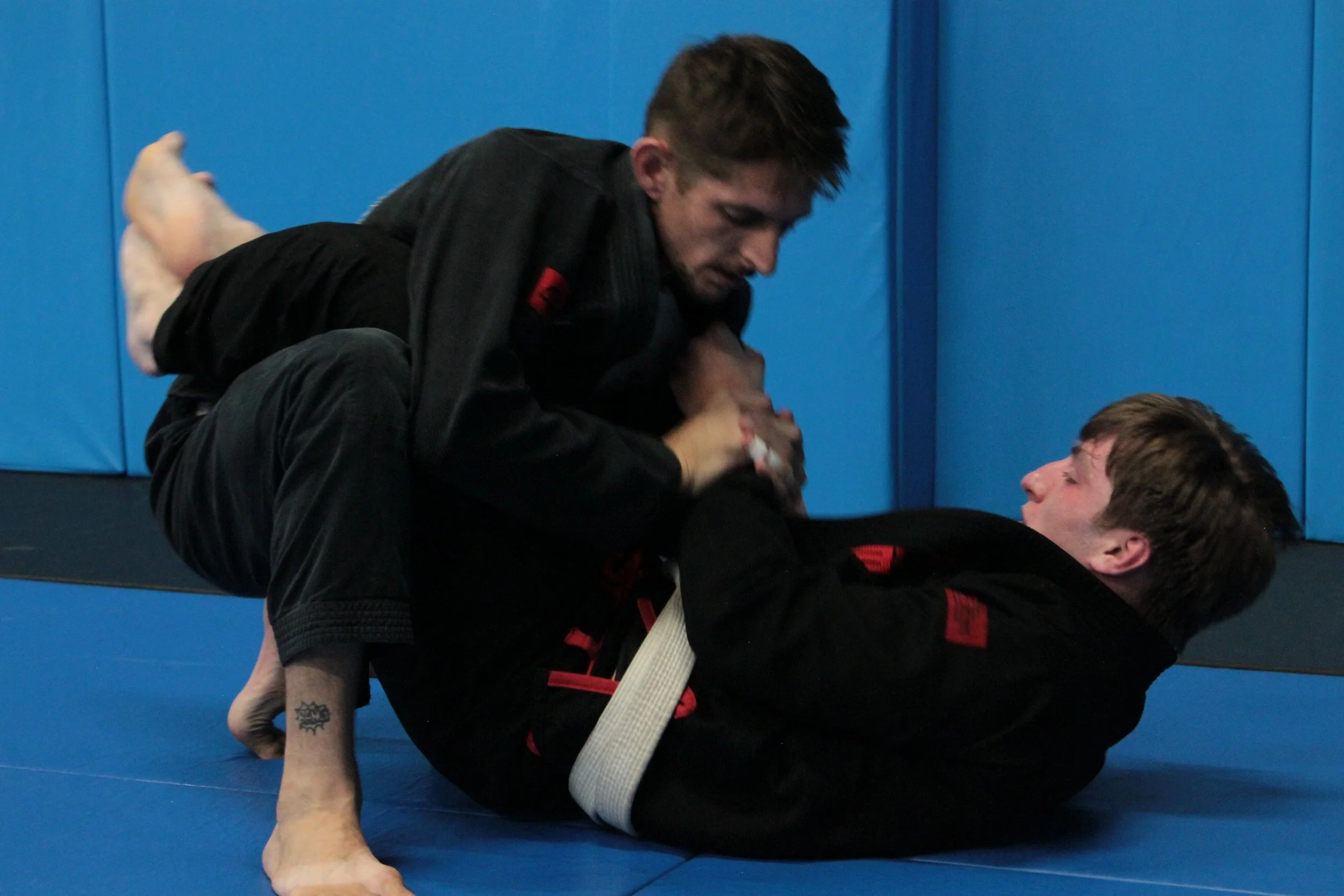 Two men practicing Brazilian Jiu-Jitsu on blue mats, with one man on top in a dominant position and the other on the bottom attempting a guard hold.