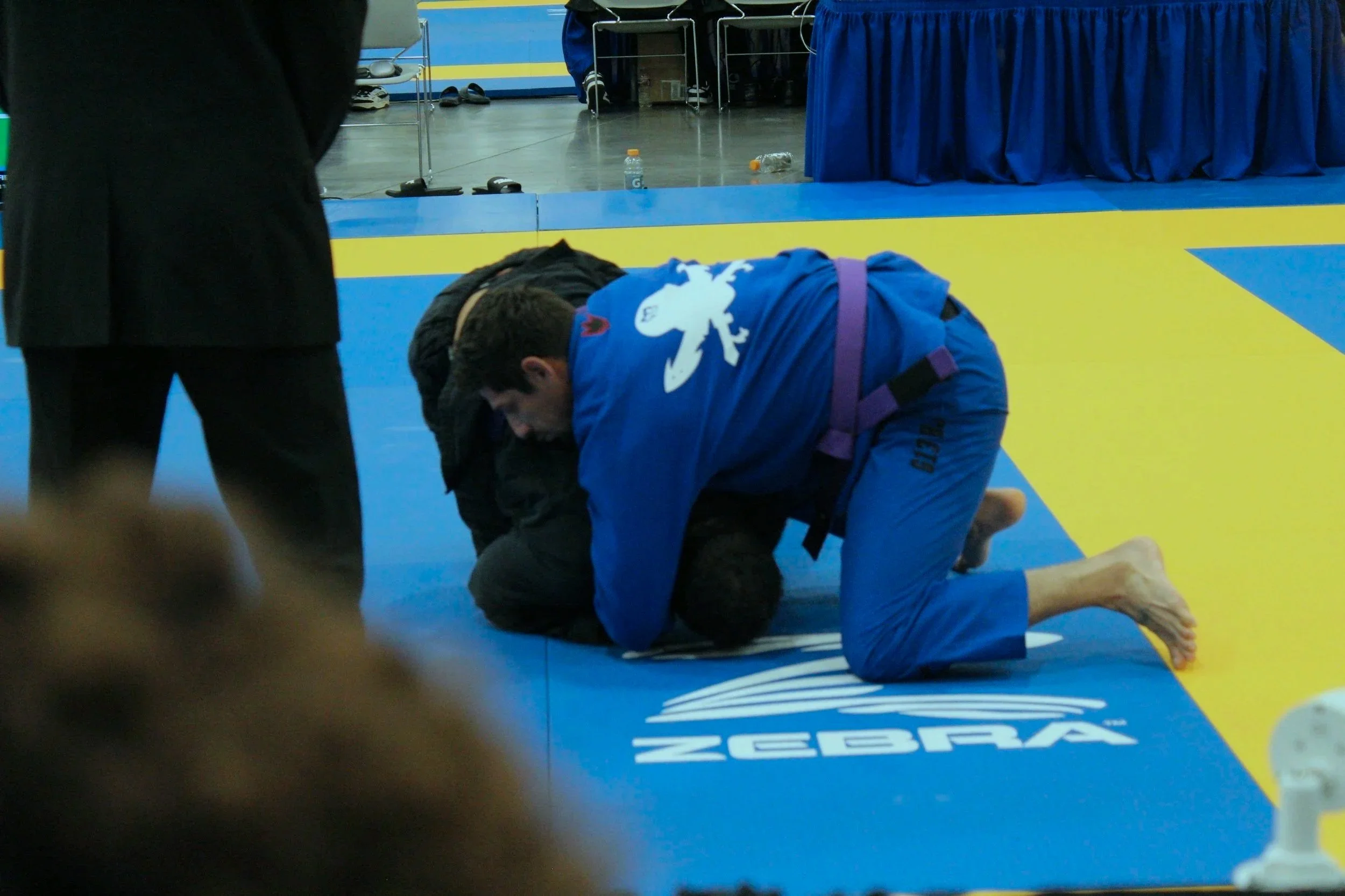 Two Brazilian jiu-jitsu practitioners in blue and black gis on a blue and yellow mat during a match, with one executing a grappling move on the other.
