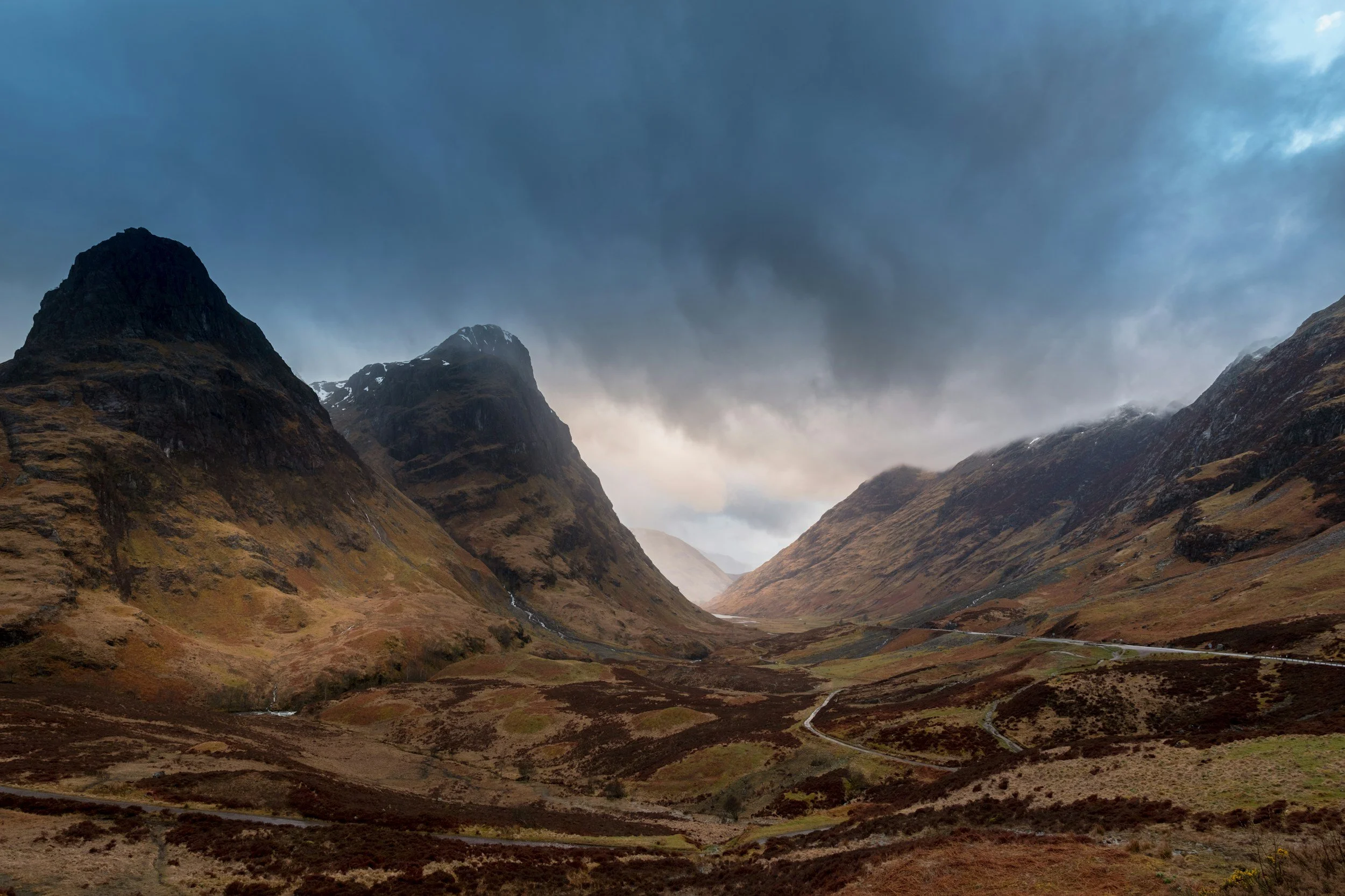 Scottish Highland mountains with dark green and brown slopes under cloudy sky, with a valley and winding road visible in the foreground.