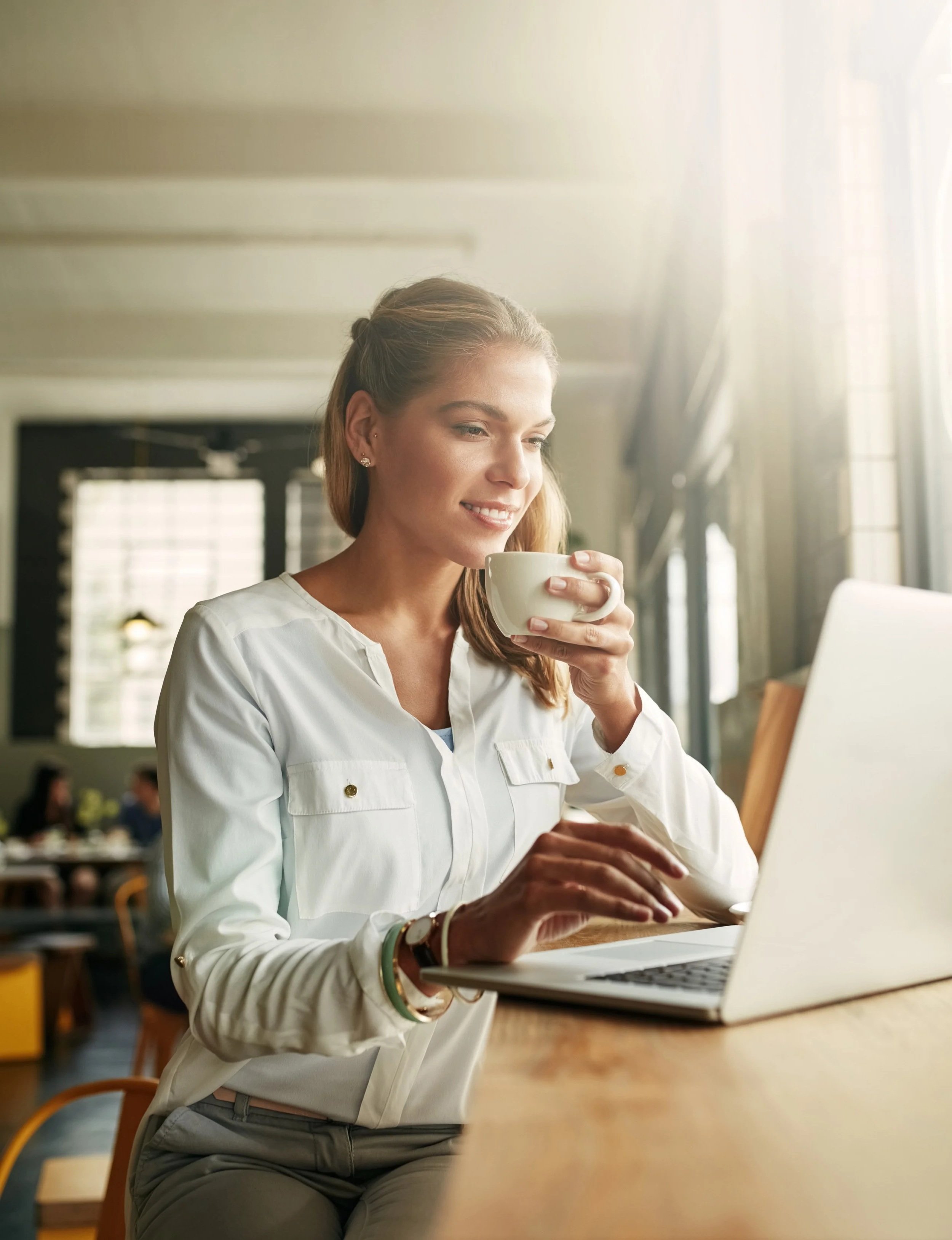 A young woman with light brown hair, wearing a white blouse, sitting at a wooden table in a cafe, holding a white coffee cup in one hand and typing on a silver laptop with the other, with sunlight streaming in from large windows behind her.