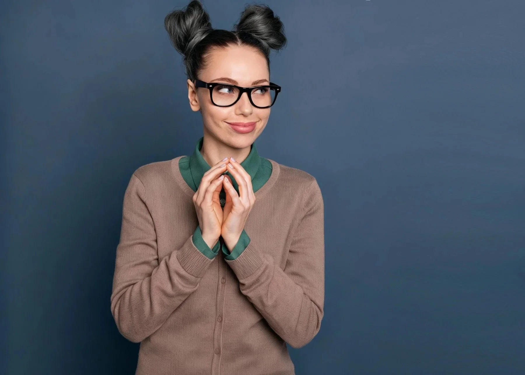A woman with glasses, styled hair, and a beige sweater with green accents, smiling with her hands clasped near her chin, standing against a plain dark blue background.
