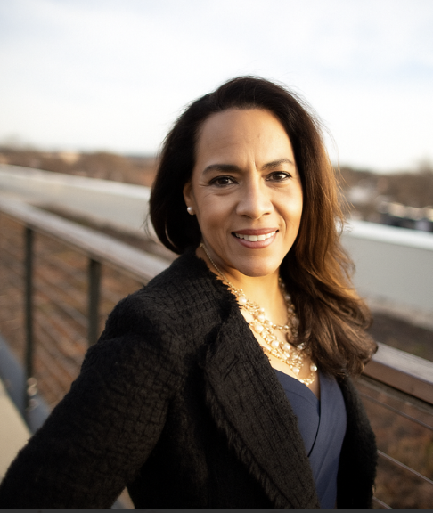 A woman with shoulder-length dark brown hair wearing a black blazer, pearl earrings, and a layered pearl necklace, standing outdoors on a balcony with a blurred landscape in the background. Social Media Management. Digital Marketing. Media Agency.