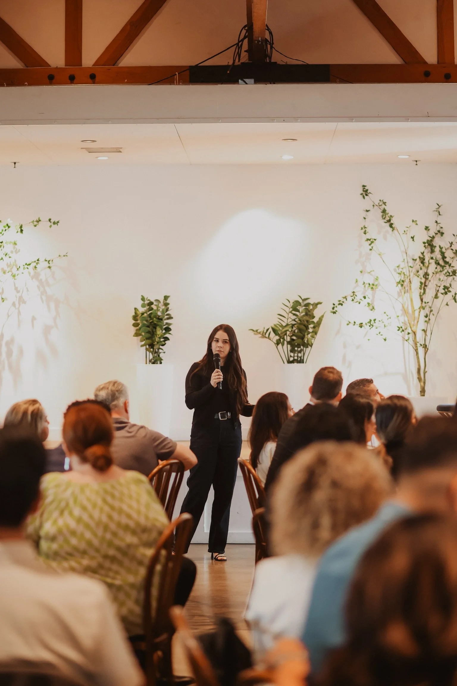 A woman in black standing and speaking into a microphone during a presentation or event, with an audience seated in front of her, in a room decorated with potted plants on a white wall. Social Media Management. Digital Marketing. Media Agency.