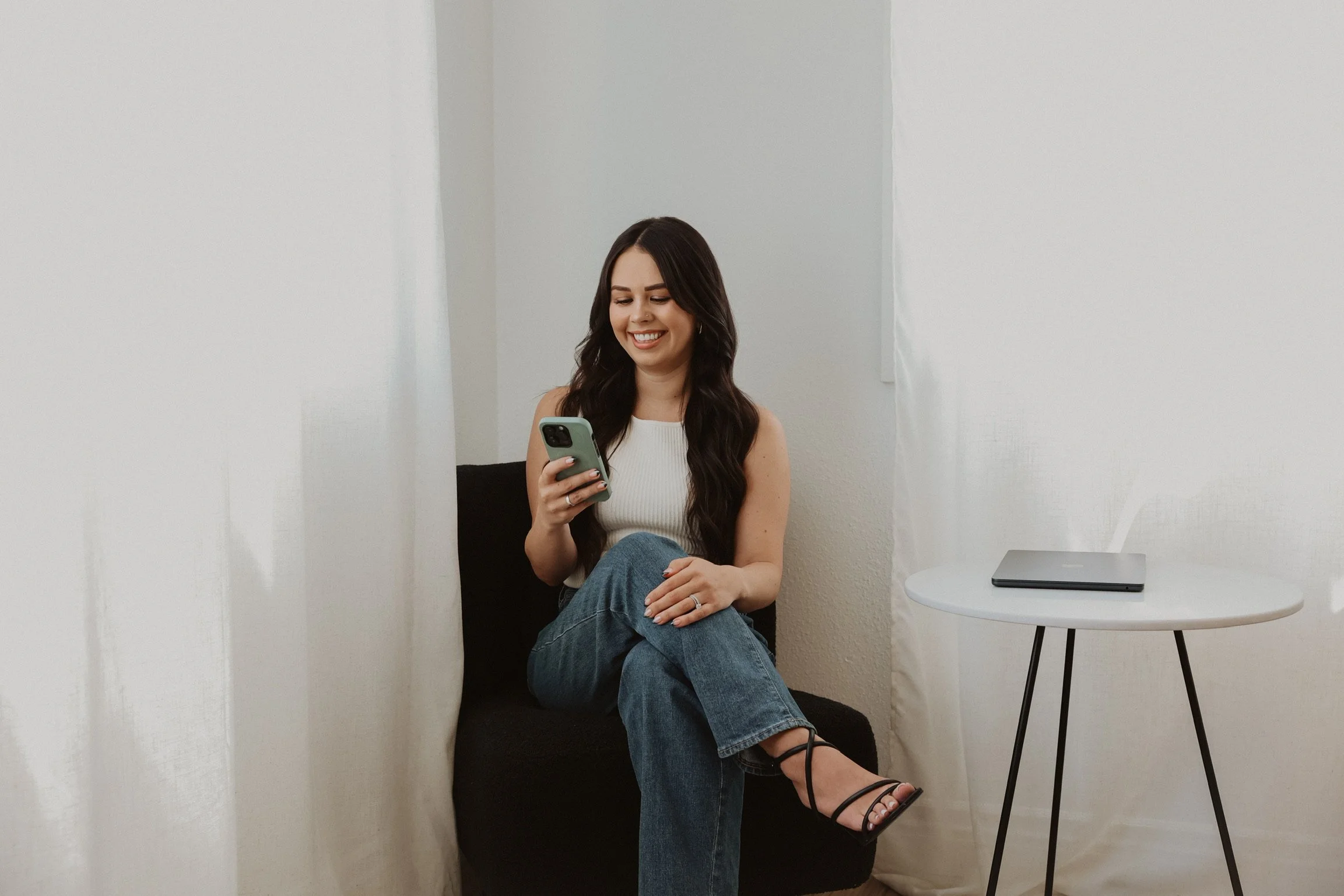 A woman with long dark hair smiling and looking at her phone while sitting on a black chair between white curtains in a bright room with a white wall and a small white round table with a closed laptop.