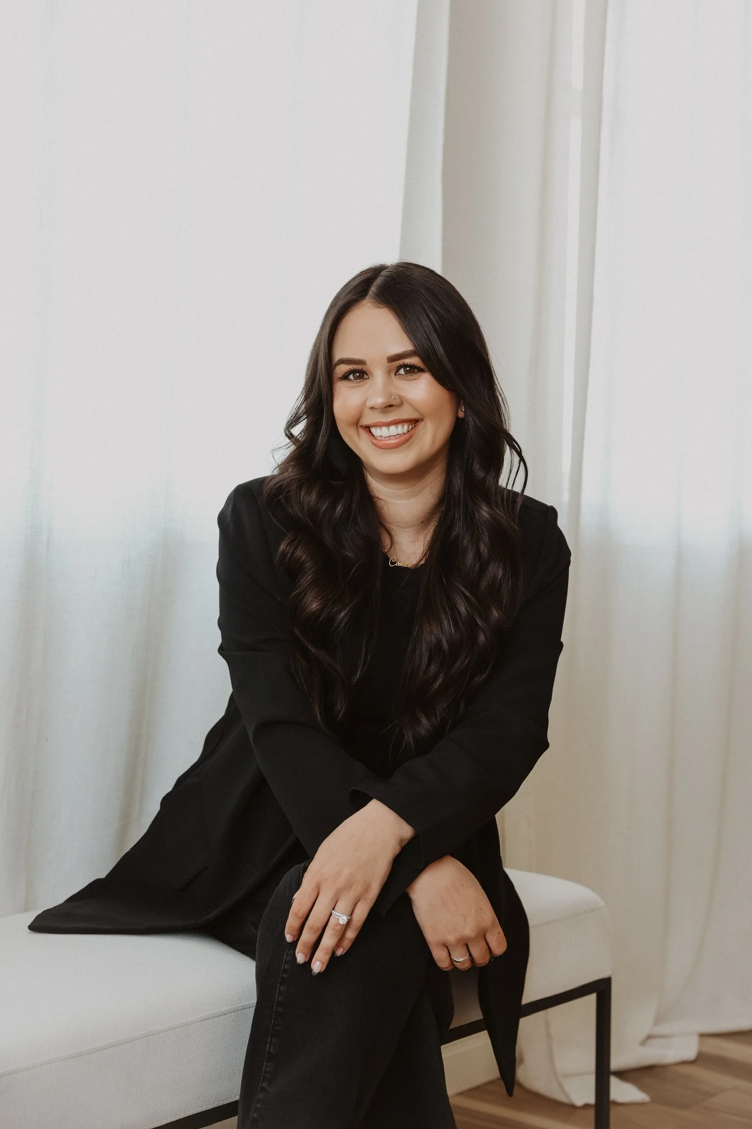 A woman with long dark wavy hair, smiling, sitting on a white bench in front of light-colored curtains, wearing a black blazer and black pants. Social Media Management. Digital Marketing. Media Agency.