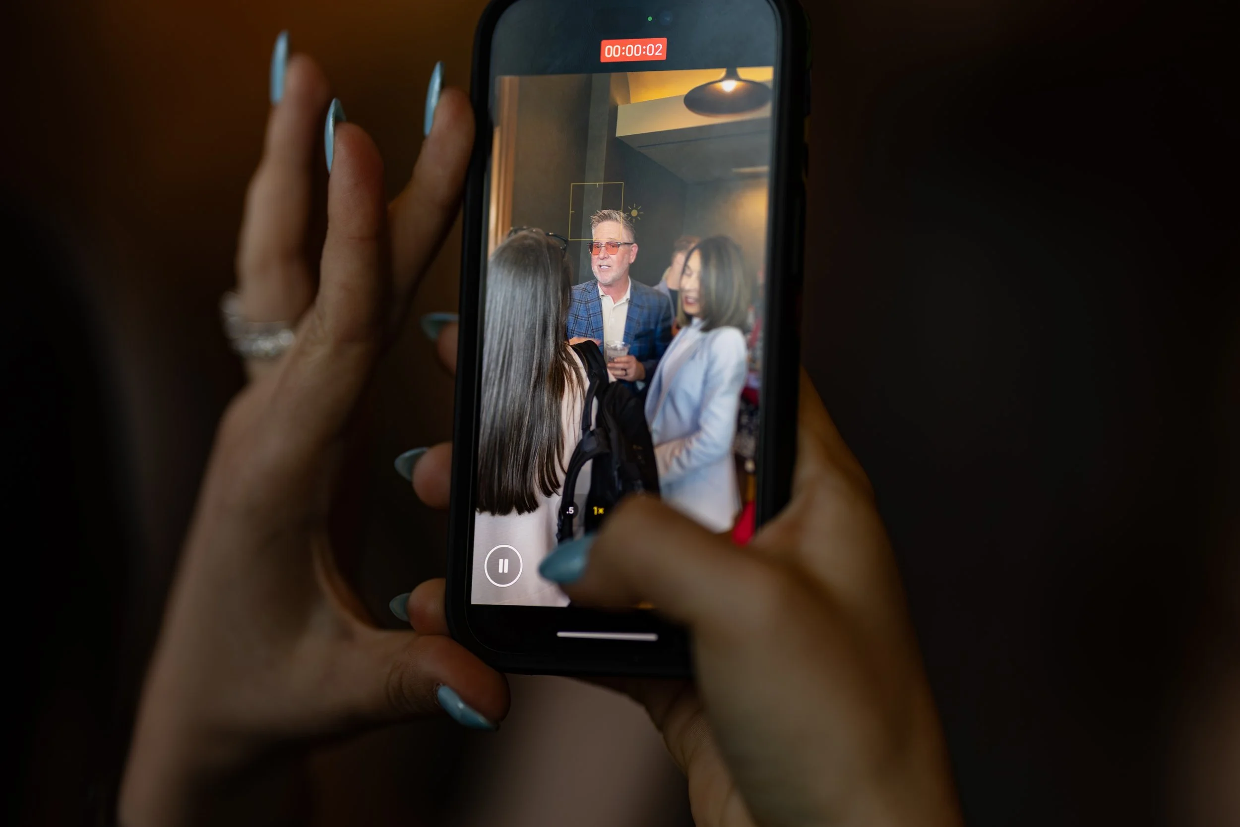 A person holding a smartphone, recording a conversation or interview with three people in a bar or restaurant. The screen shows a man with glasses and two women, all engaged in conversation. Social Media Management. Digital Marketing. Media Agency.