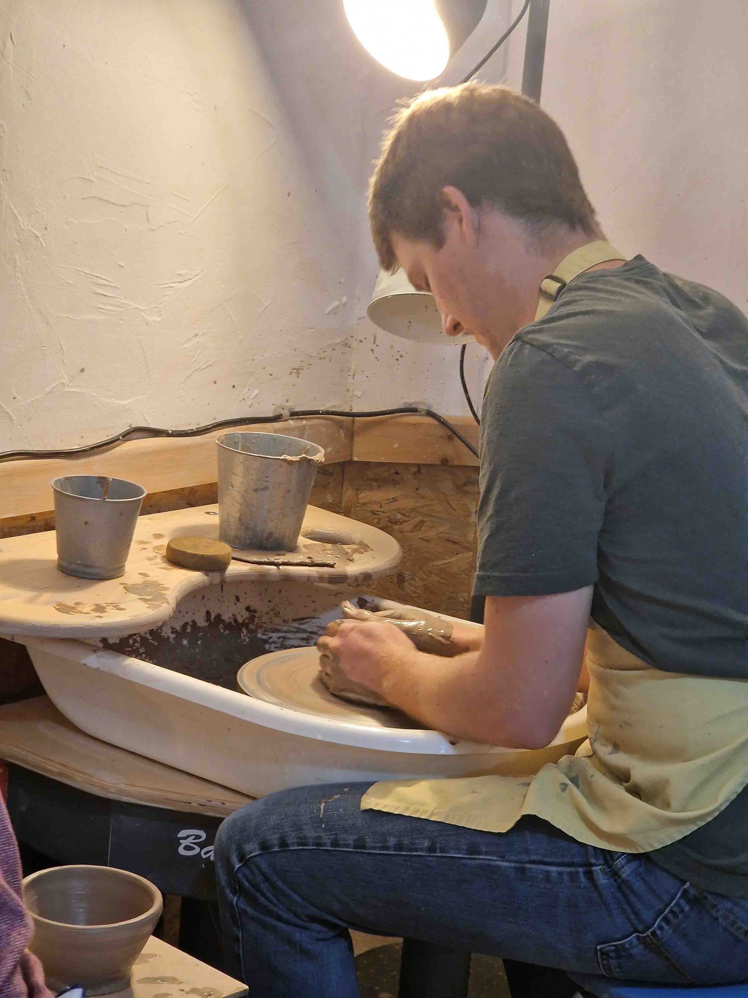 Young man shaping clay on a pottery wheel in a studio, with pottery tools and containers nearby.