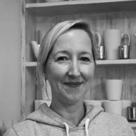 Black-and-white photo of a woman with short hair smiling, standing in front of a wooden shelf with various mugs and cups.
