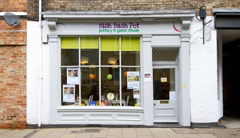 The storefront of BiSh Bash Pot pottery and glass studio, featuring a large display window and a white door, with various posters and decorations inside visible through the window.
