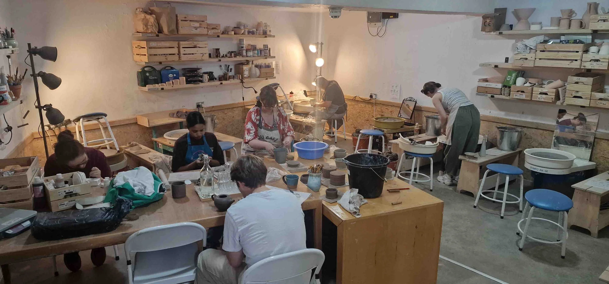 People in a pottery studio working on pottery projects, surrounded by pottery tools, shelves with supplies, and a large work table.