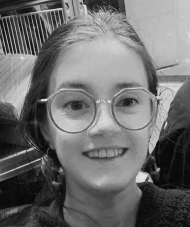 A young girl with glasses smiling at the camera, with a fence and outdoor seating area in the background.