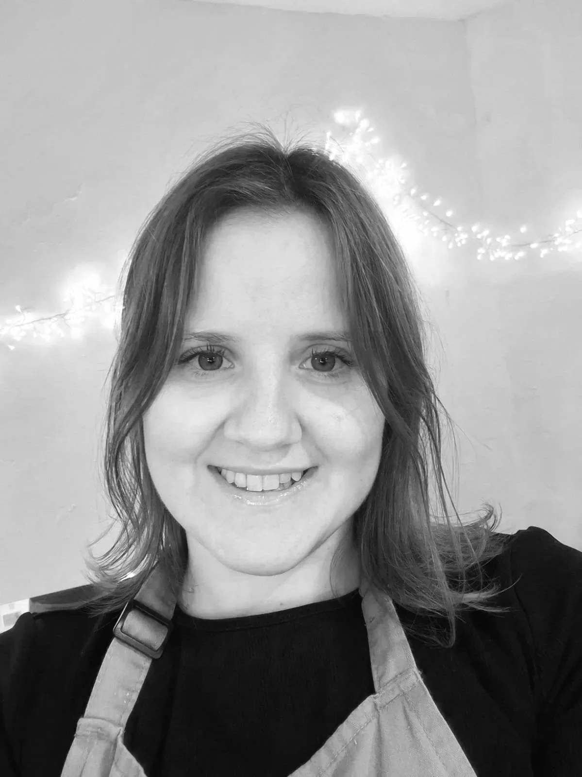 A woman with light skin, shoulder-length hair, smiling, wearing a black shirt and apron, with string lights in the background.