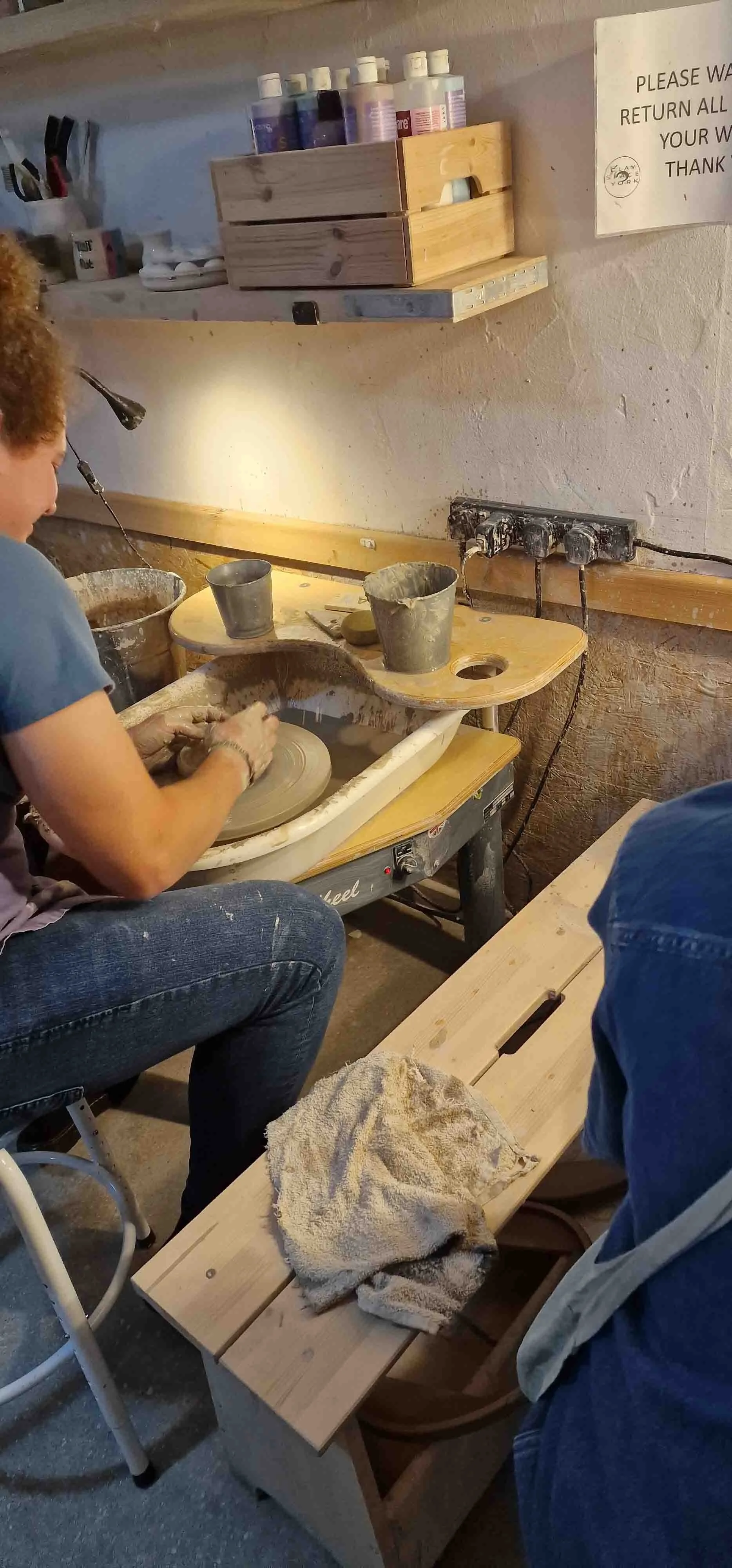 A person working on a pottery wheel, shaping clay into a vessel, with pottery tools and buckets nearby in a pottery studio.