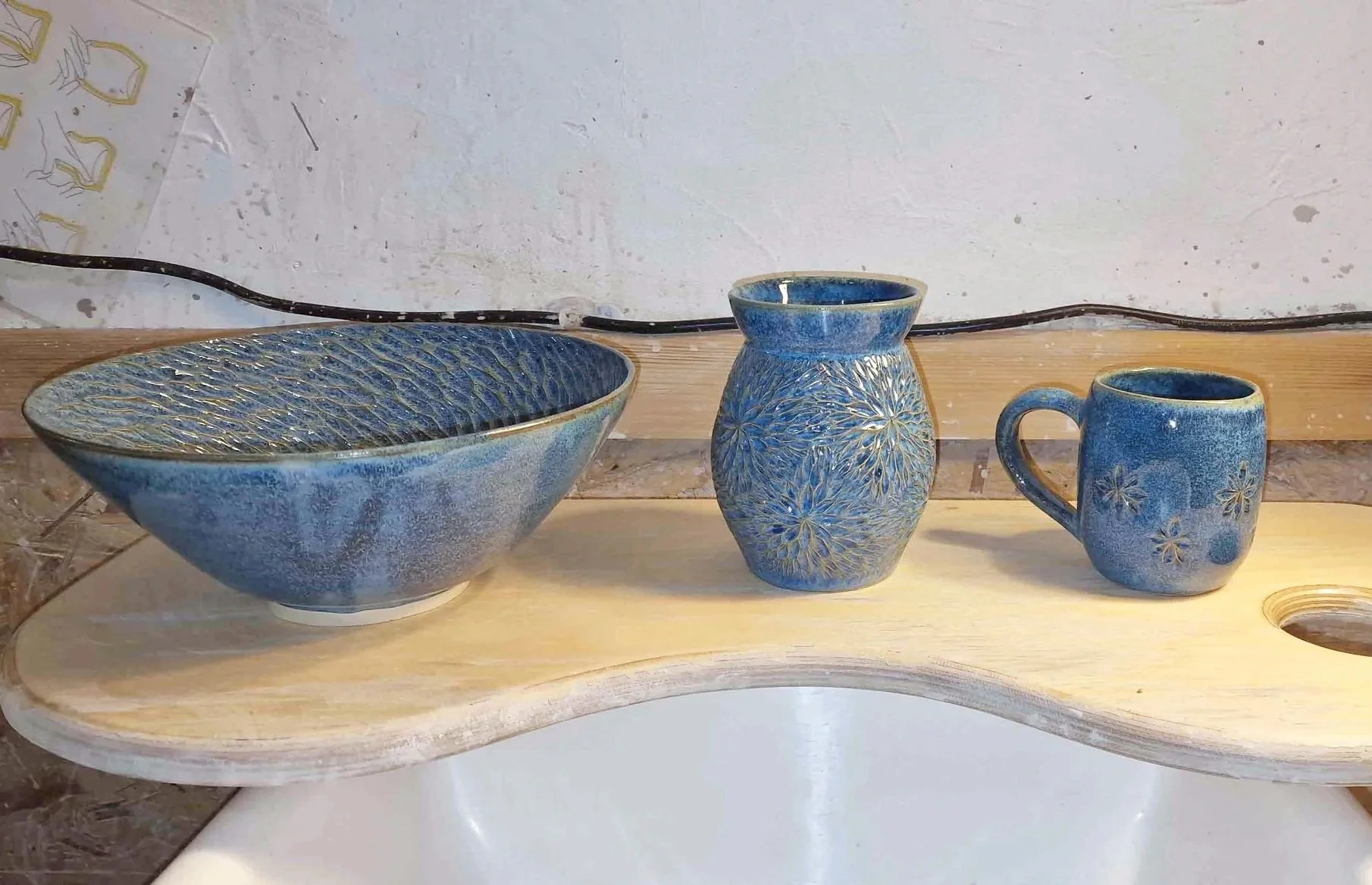 Three pieces of blue ceramic pottery on a wooden shelf: a large bowl, a small vase, and a mug, all with intricate floral designs or textures.