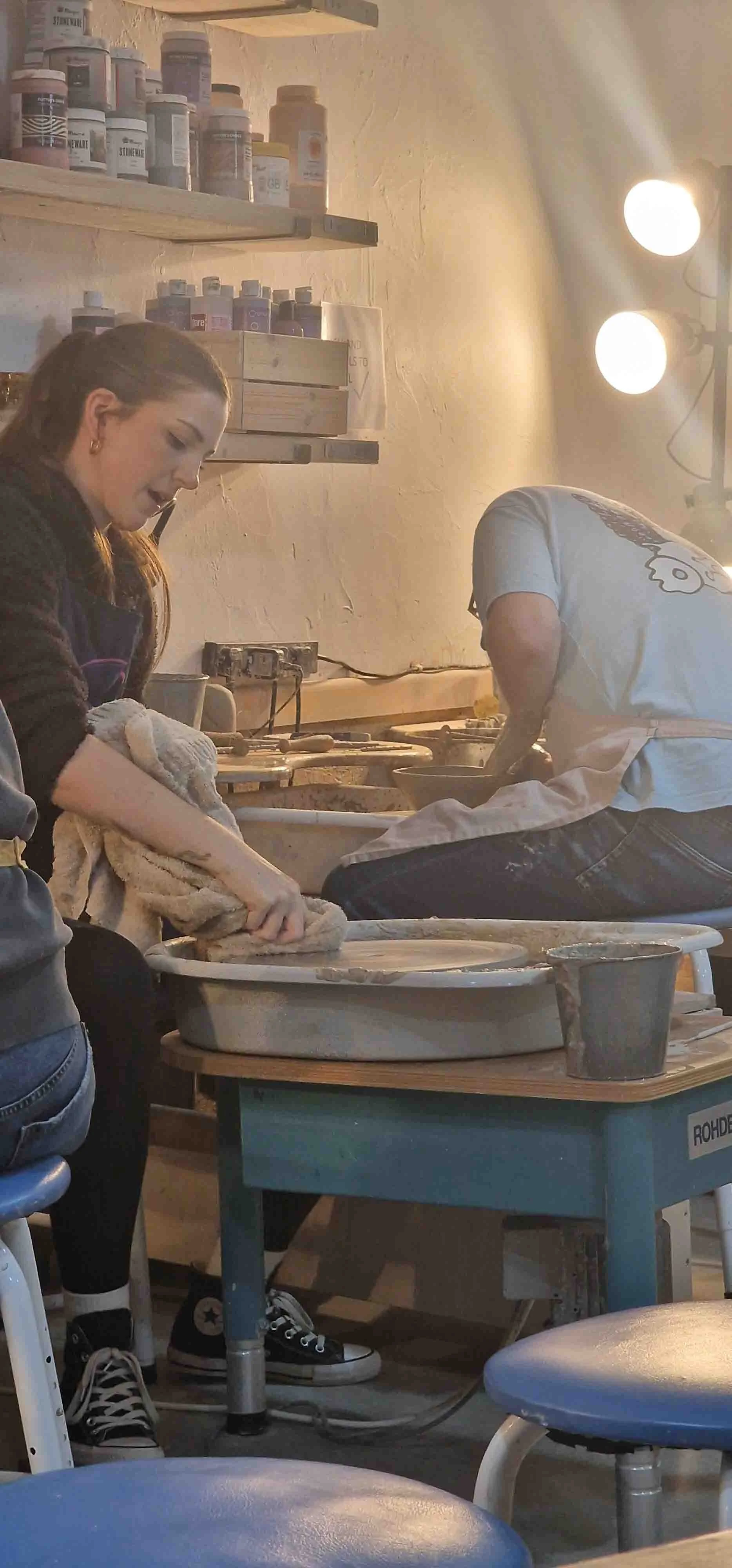 A young woman working at a pottery wheel in a ceramics studio, with another person bent over their work in the background, shelves with containers of paint or glaze visible on the wall, and bright studio lights overhead.