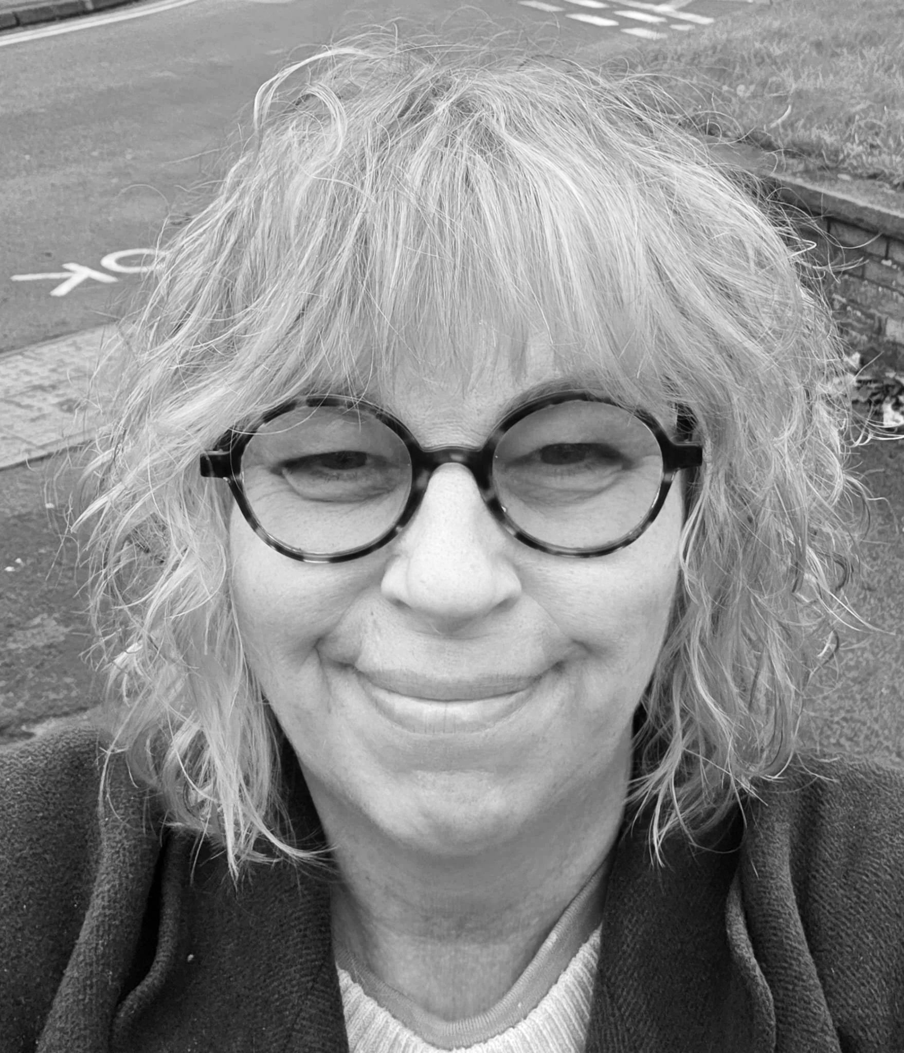 A woman with curly, light-colored hair and round glasses smiling at the camera in a black-and-white photo.