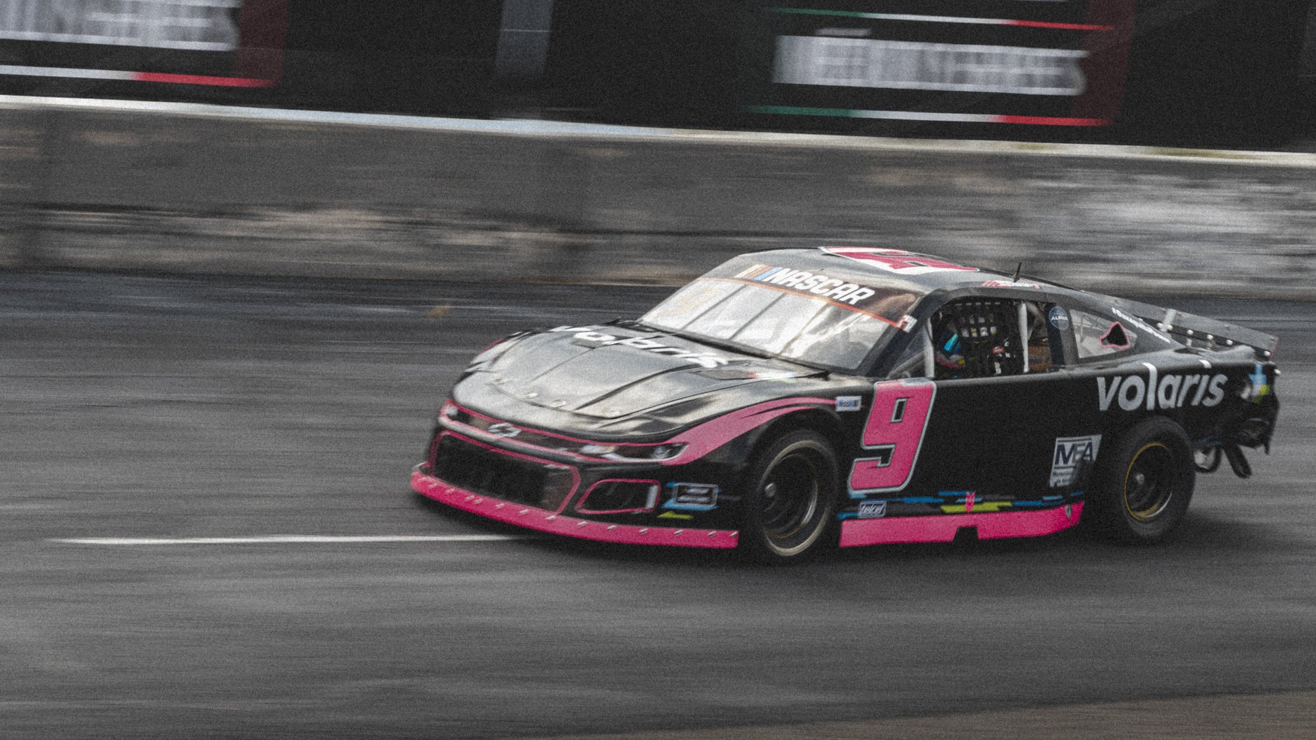 Automóvil de carreras en pista de NASCAR, color negro con detalles en rosa y patrocinadores en los laterales, número 9.