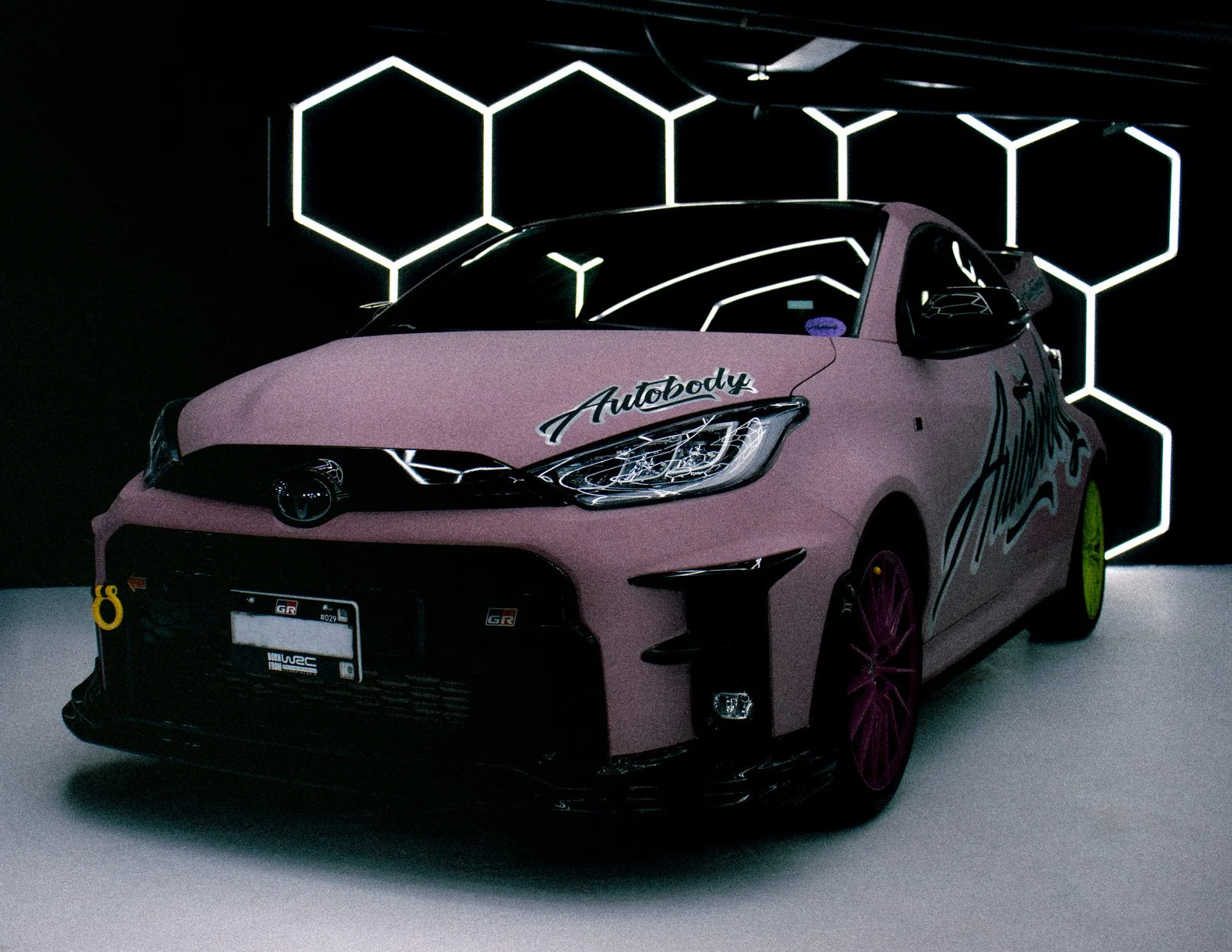 Auto compacto de color rosa con detalles en negro y palabras decorativas, exhibido en un entorno oscuro con decoraciones de luces en forma de hexágonos.