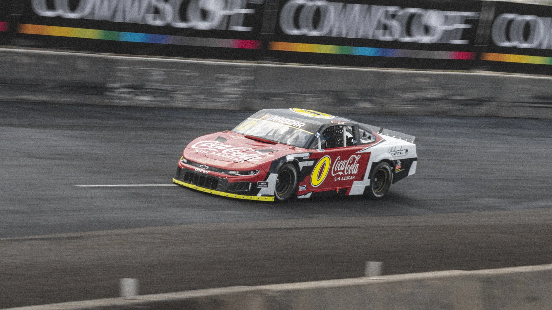 Coche de carreras con anuncios de Coca-Cola y Q en color rojo, blanco, negro y amarillo, en una pista de carreras.