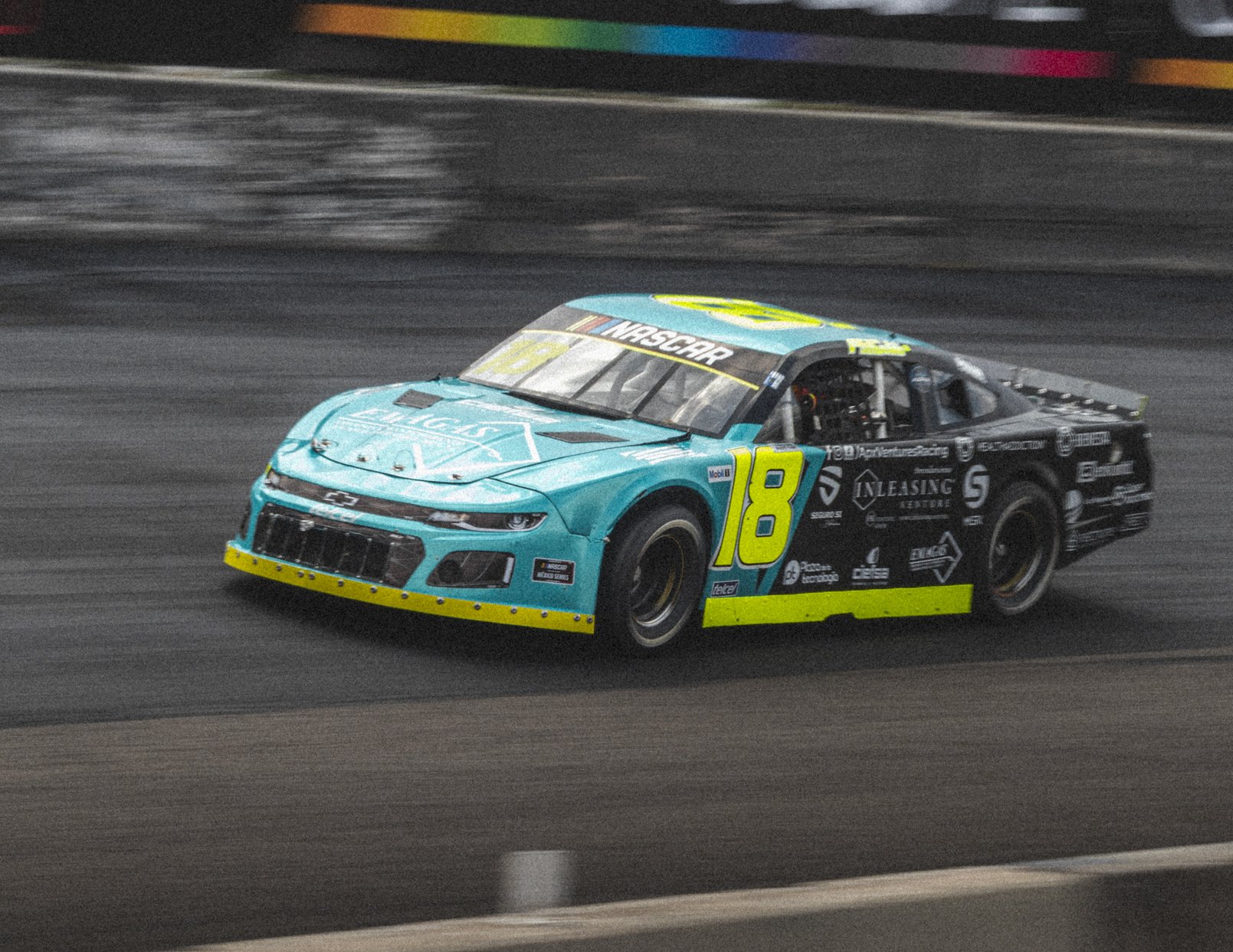Coche de carreras azul y negro con el número 18 en amarillo, en una pista de carreras de NASCAR.