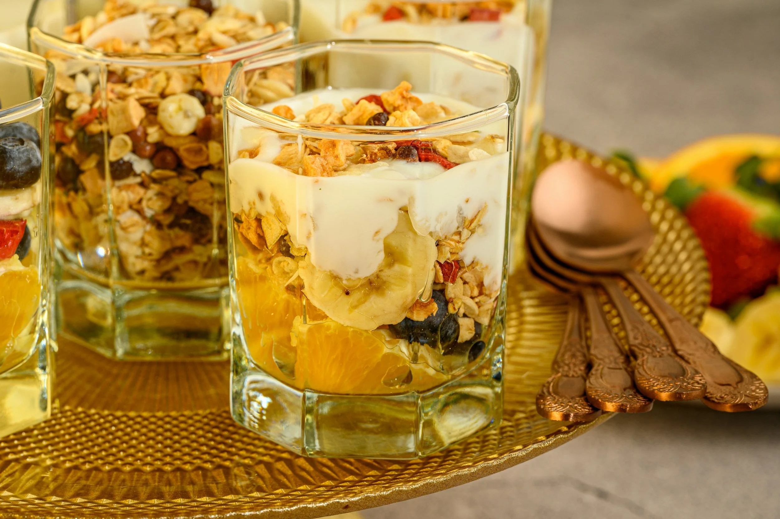 Close-up of a glass containing parfait with granola, yogurt, and fresh fruit, placed on a gold-colored textured tray.