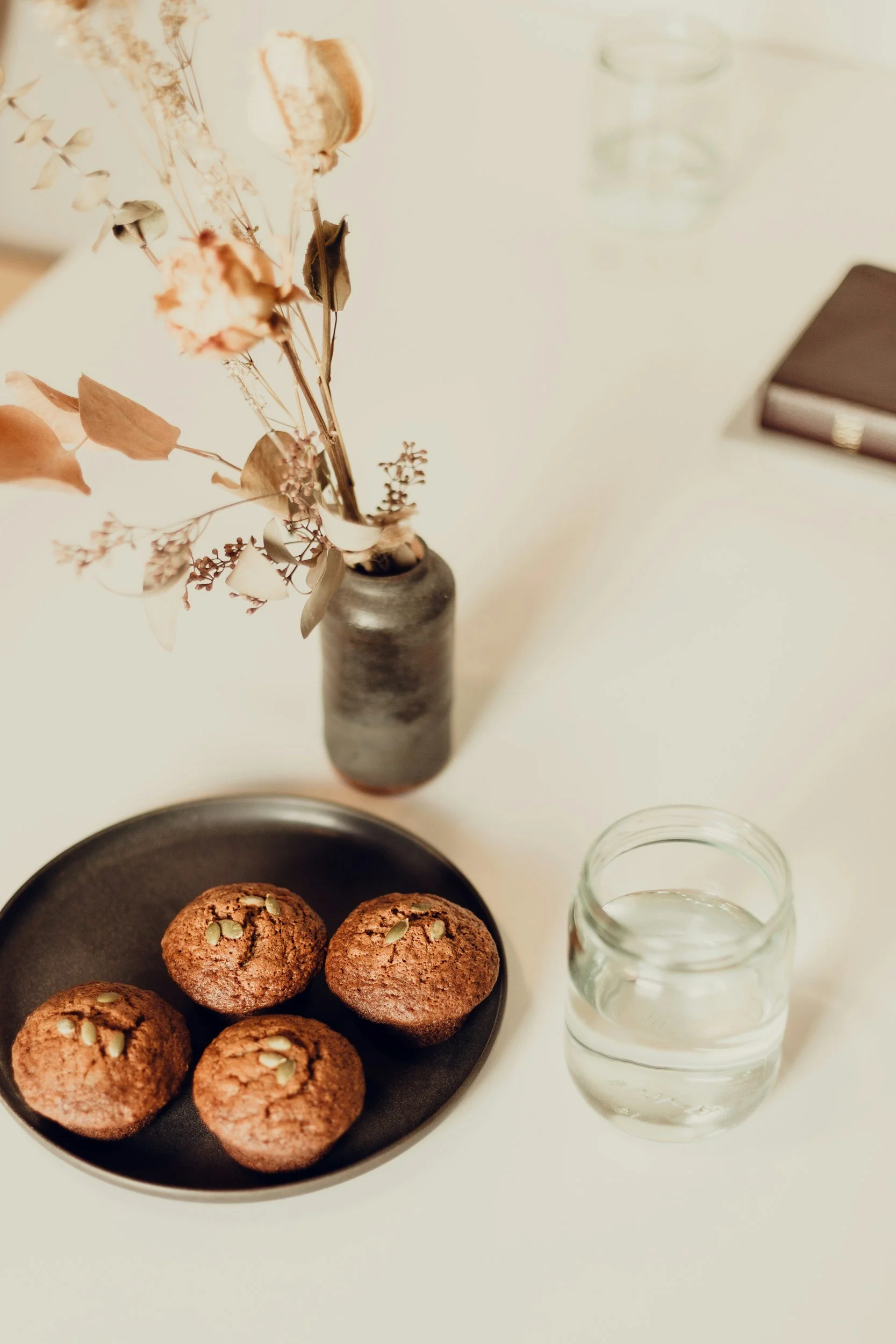 A table with a black plate of six chocolate cookies topped with pumpkin seeds, a gray vase with dried flowers, a clear glass of water, an empty glass, and a closed brown notebook.