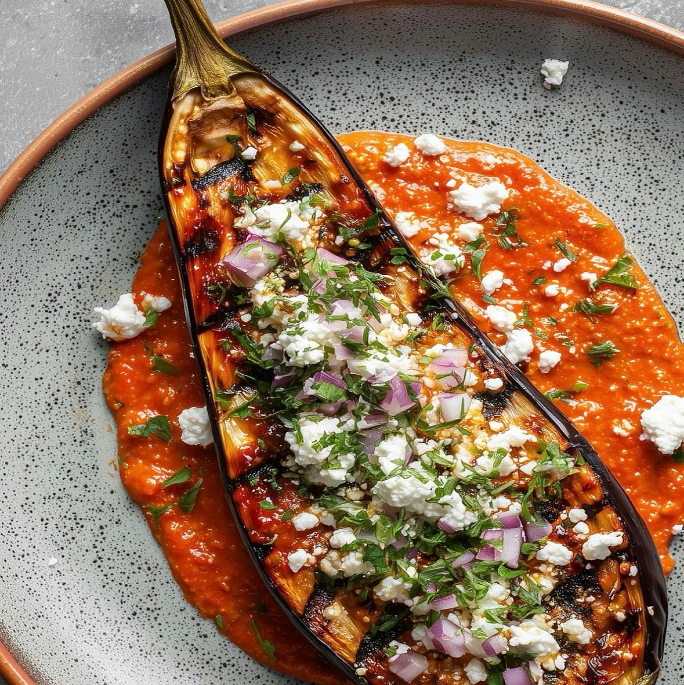 A ceramic bowl with a dotted gray interior contains a roasted or grilled eggplant half topped with sauce, chopped red onion, herbs, and dollops of white cheese, with additional sauce spread on the plate.