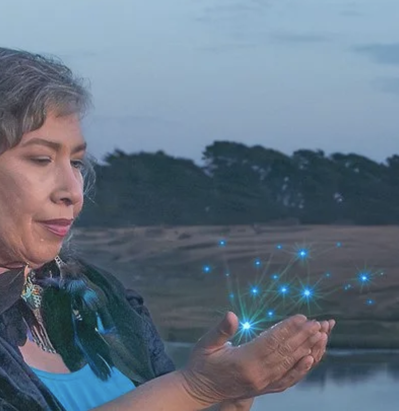 A woman with gray hair holding her hands close to her face, with glowing blue digital points and lines emerging from her hands, set against a natural landscape background with trees and a cloudy sky.