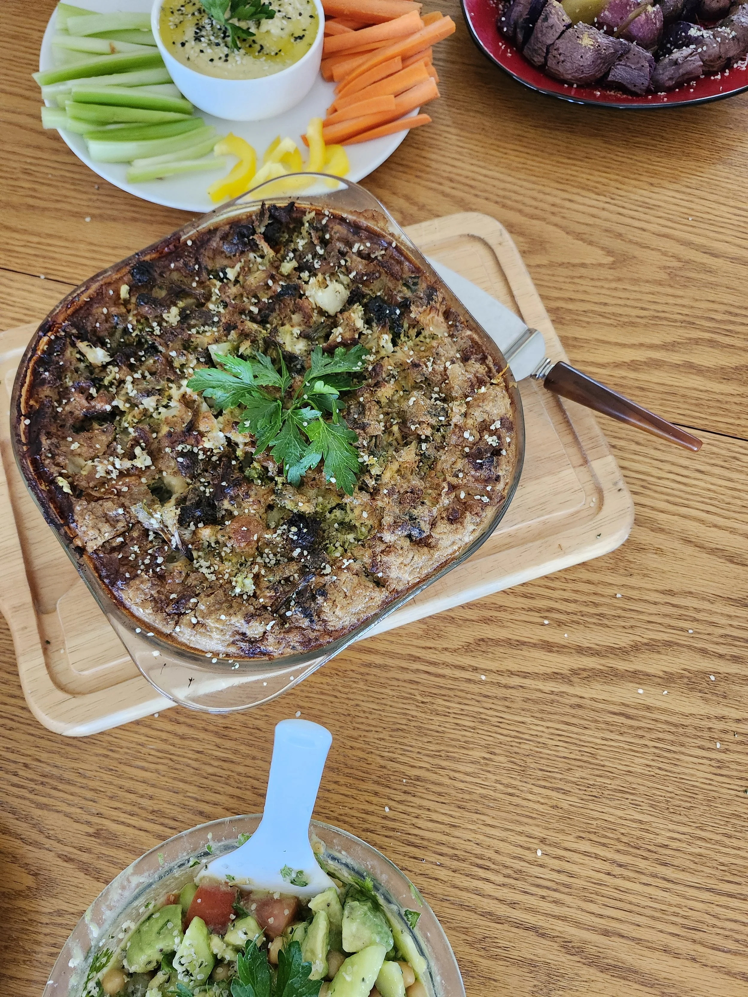 A wooden table with a glass dish of salad containing cucumbers, tomatoes, chickpeas, and parsley, a baked casserole topped with herbs, a white bowl of sauce, a plate of sliced carrots with yellow peppers, and a plate of cooked beef cubes.