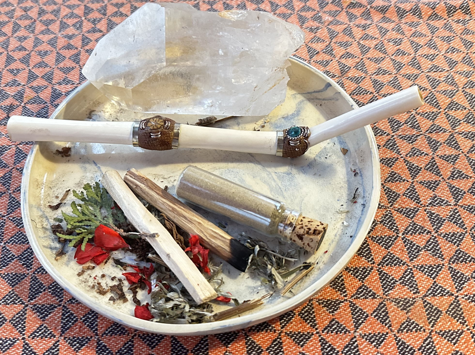 A round metal tray containing a large clear quartz crystal and medicinal tools placed on an orange and black patterned cloth.
