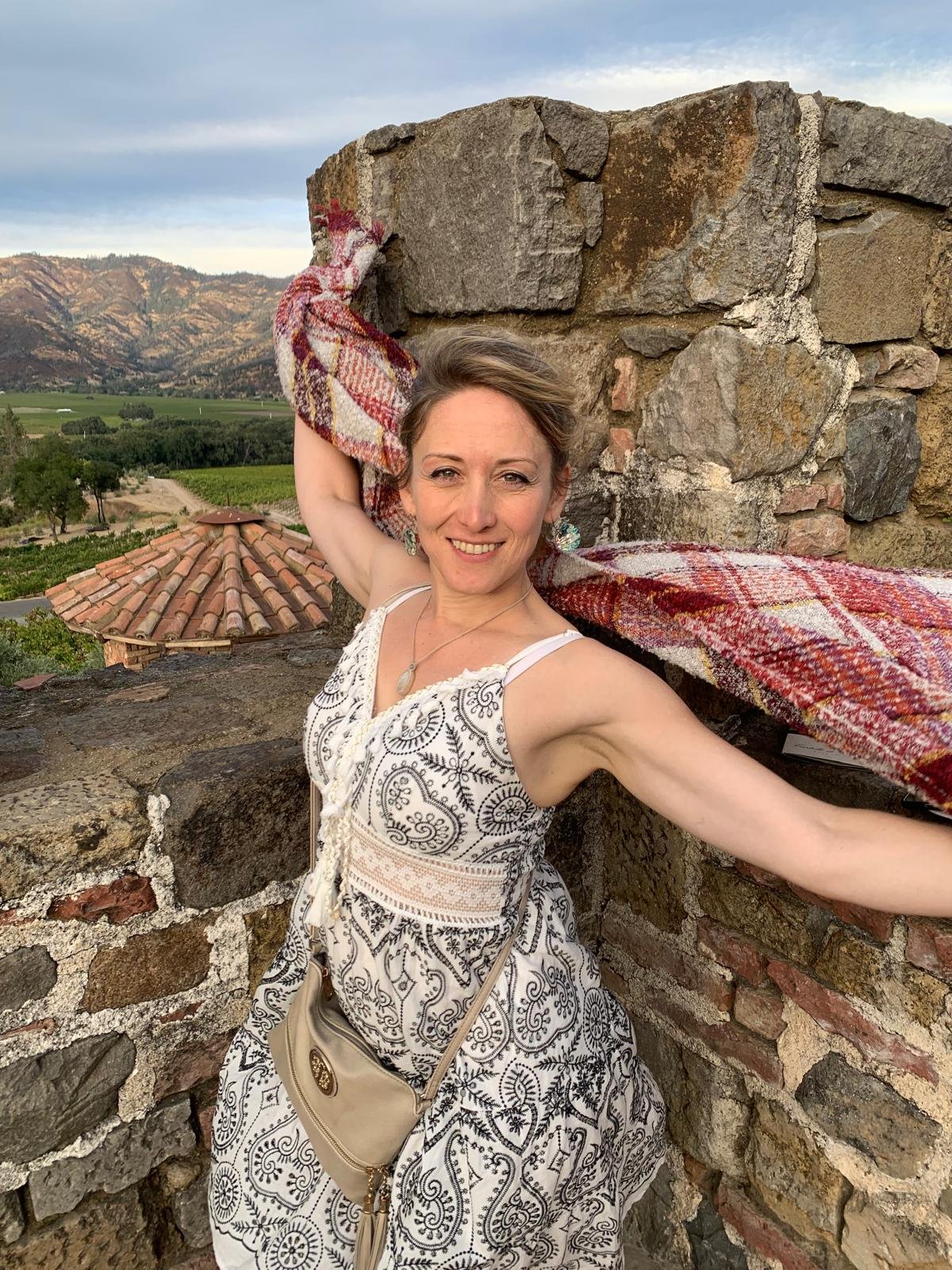 A smiling woman in a patterned white dress stands on a stone balcony with her arms outstretched. She holds a red and beige plaid scarf over her head, and there is a scenic landscape with mountains, greenery, and a tiled roof in the background.