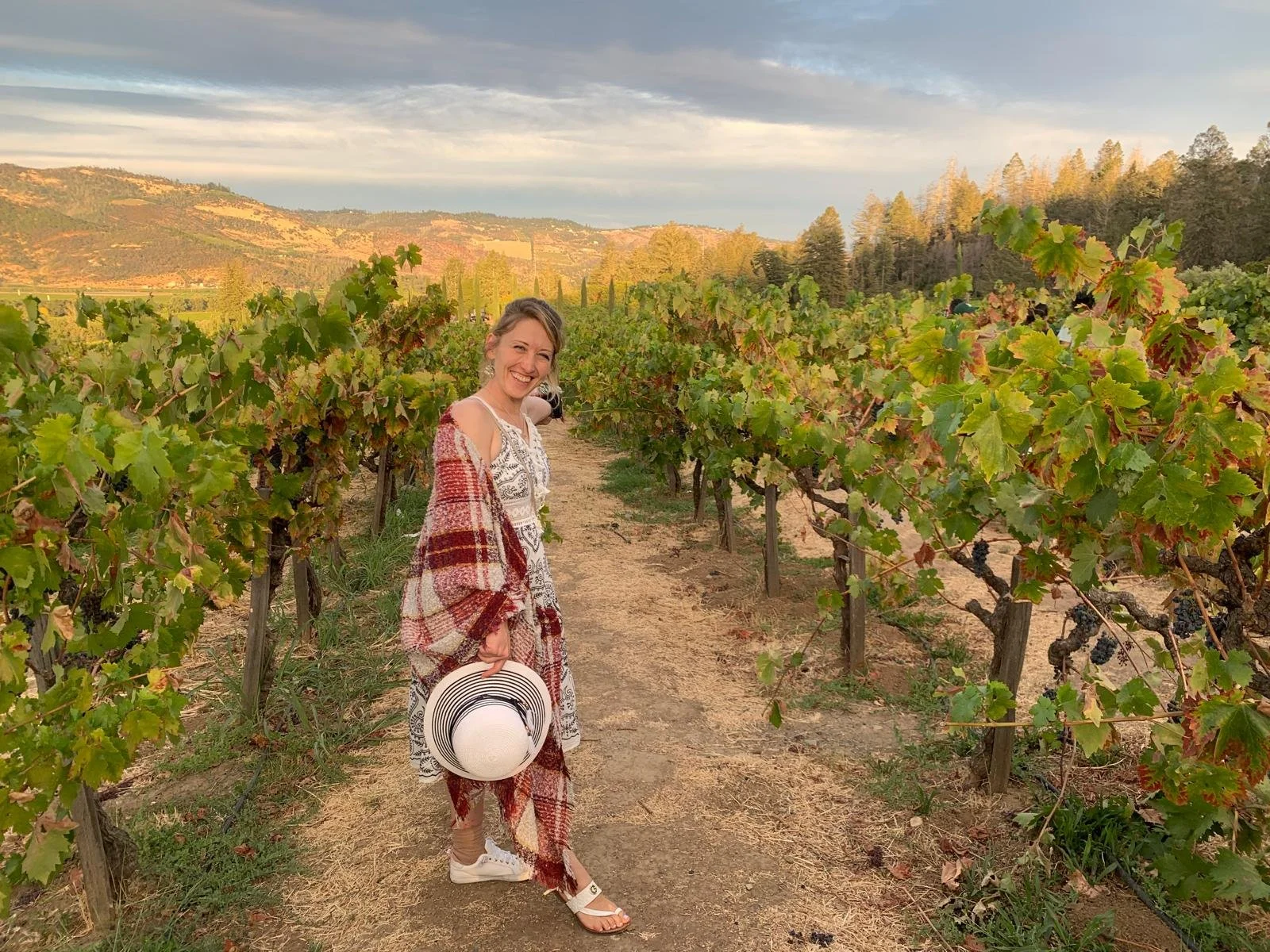 A woman smiling and holding a white sun hat with black stripes, standing in a vineyard during sunset, with green vines and a scenic landscape of rolling hills and trees in the background.