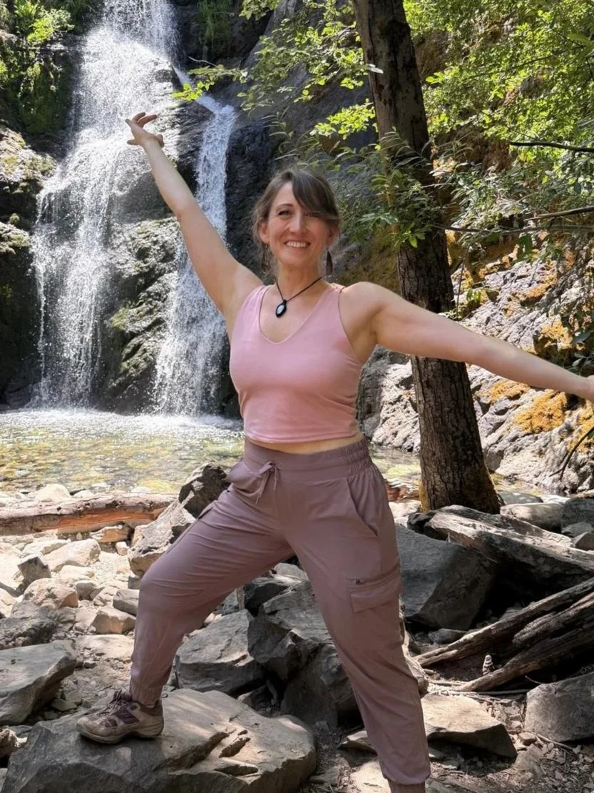 A woman smiling and posing with arms outstretched in front of a waterfall and wooded area.