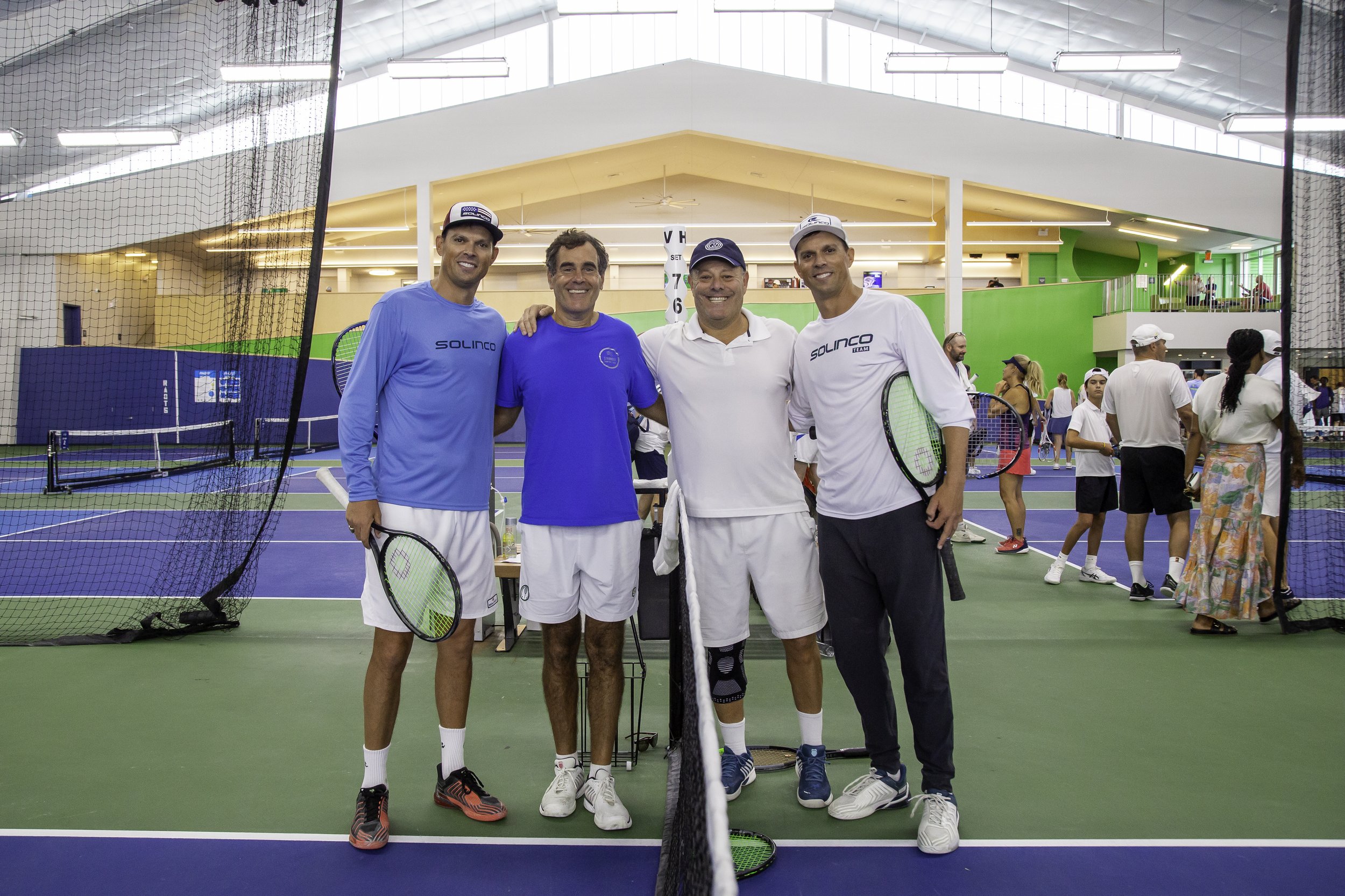 Four men standing together on an indoor tennis court. They are smiling and holding tennis rackets.