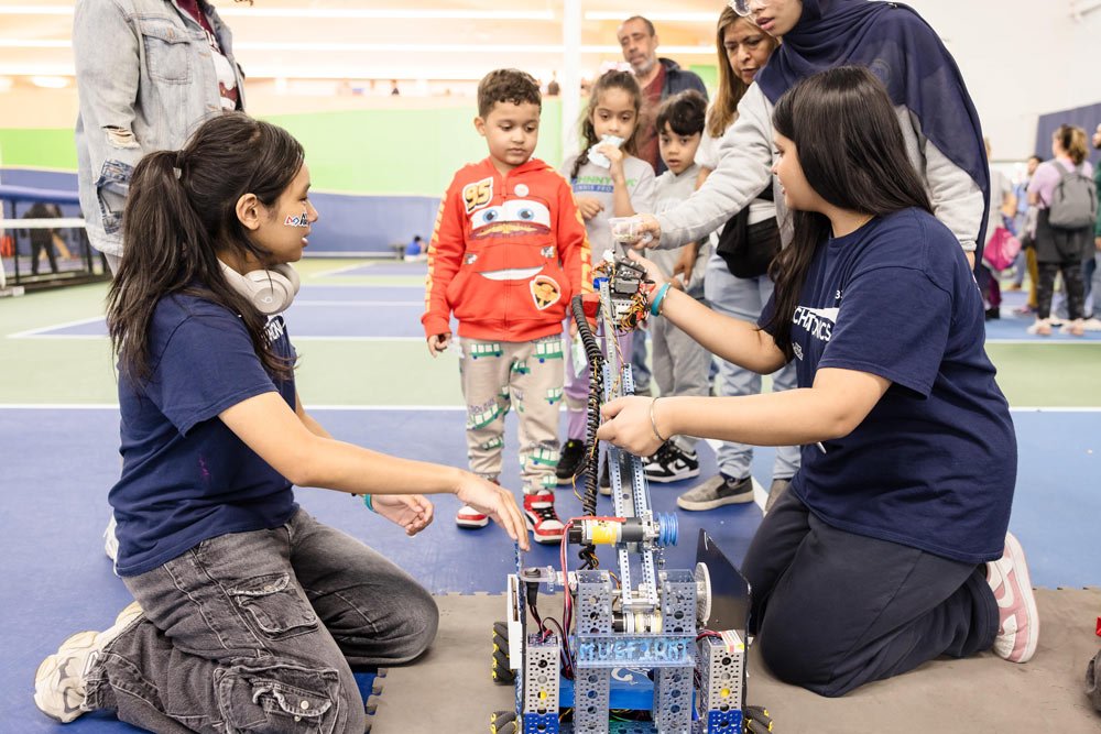 Children and adults gathered around a robotics project on a mat inside a gymnasium.