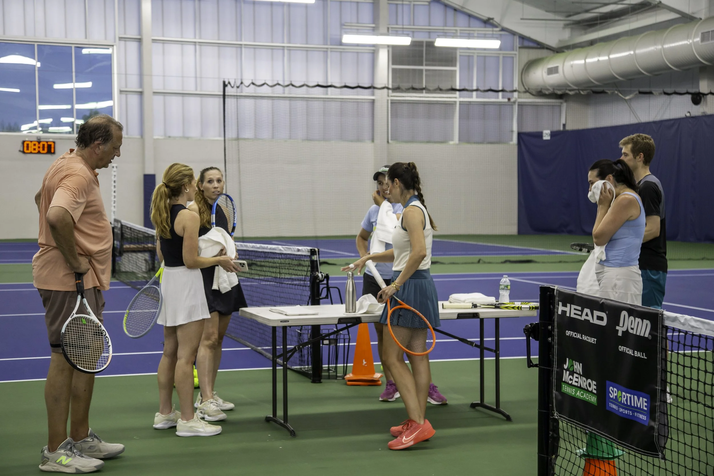 Tennis players and coach inside an indoor tennis court during a practice or meeting.