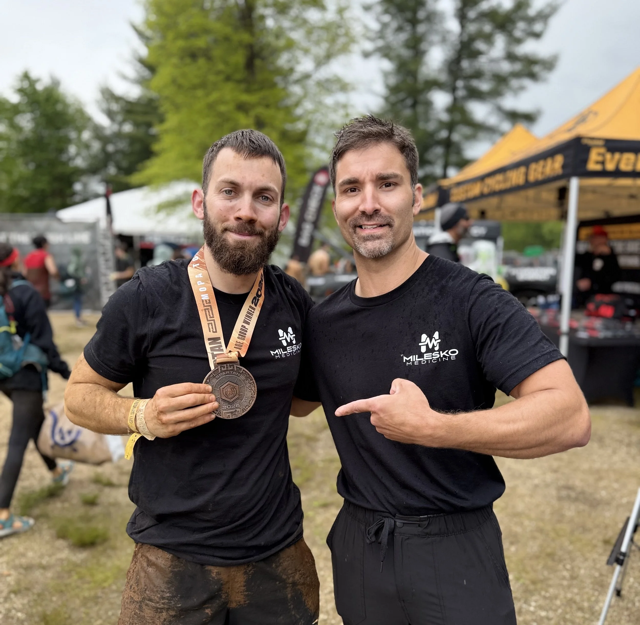 Two men wearing black shirts, both with medals around their necks, standing outdoors at a race event with a crowd and green trees in the background.