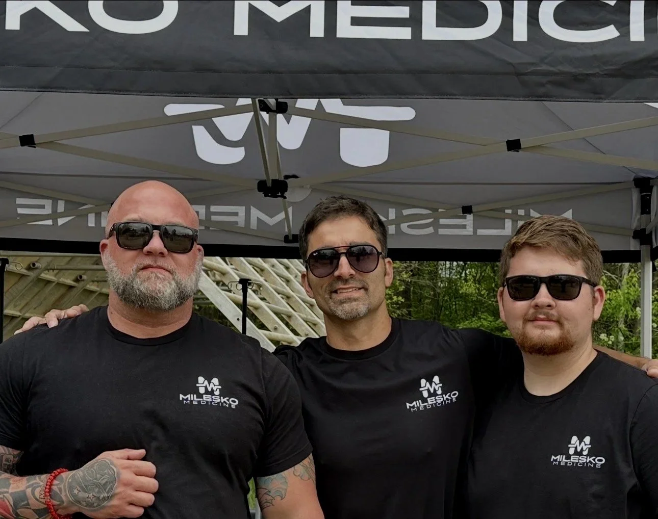 Three men standing together under a tent, wearing black T-shirts with 'Milesko Medicine' logo and sunglasses.