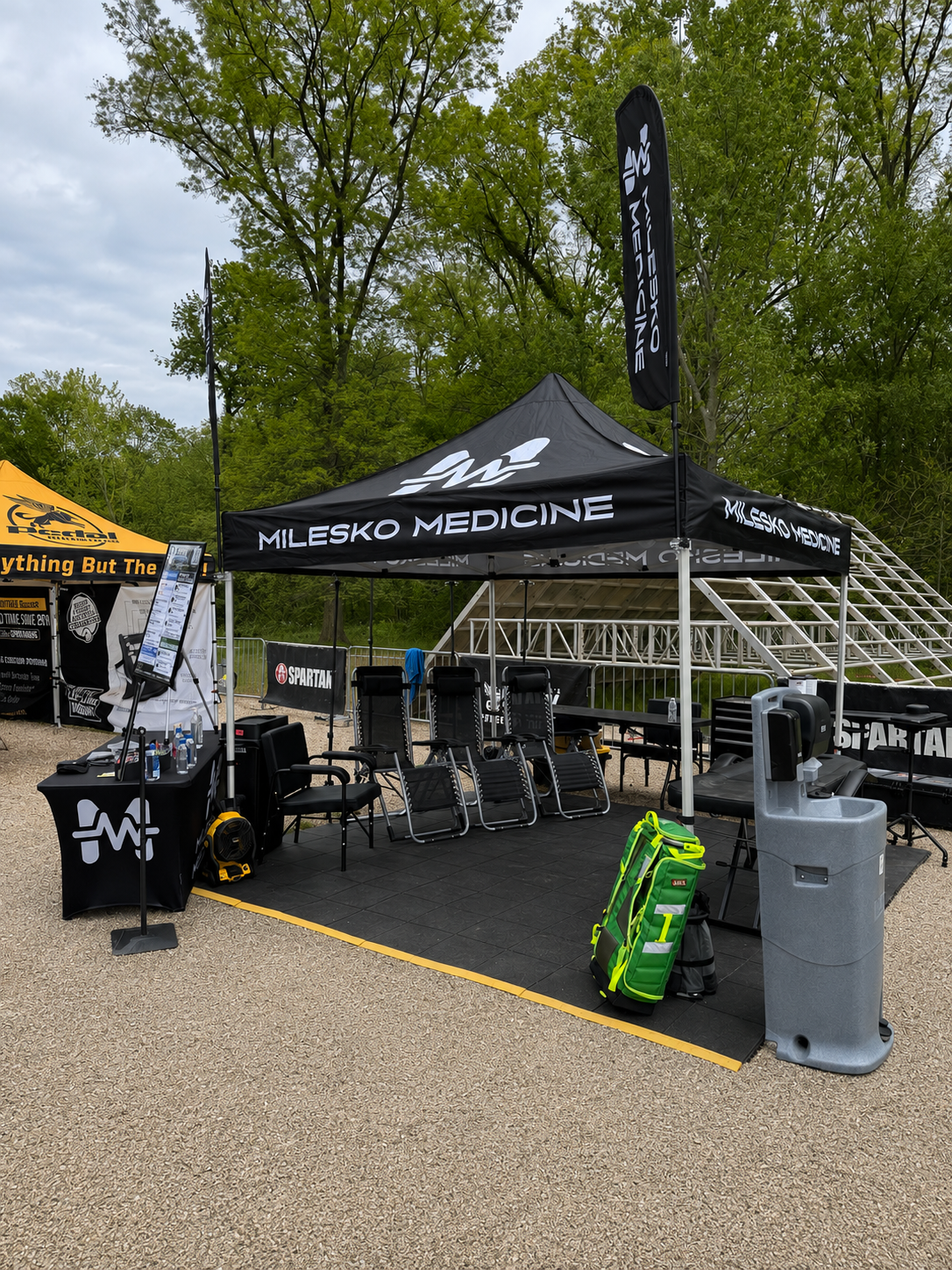 Outdoor setup with a black tent and chairs, medical supplies, and a water fountain, surrounded by trees.
